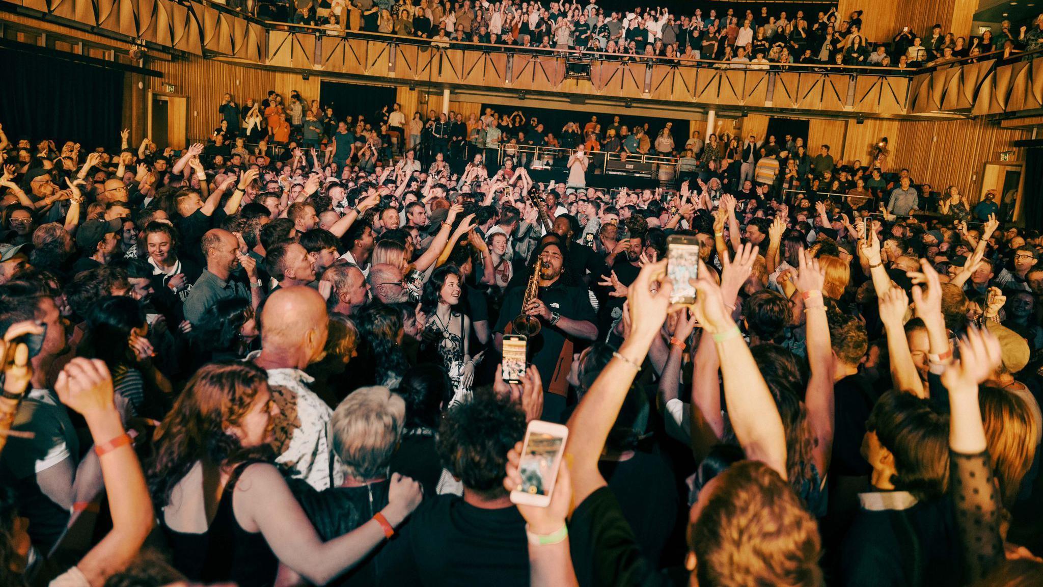 A large crowd at Bristol Beacon gather round musicians from Ezra Collective as the play in the venue's auditorium. Many of the audience are filming the musicians on their phones