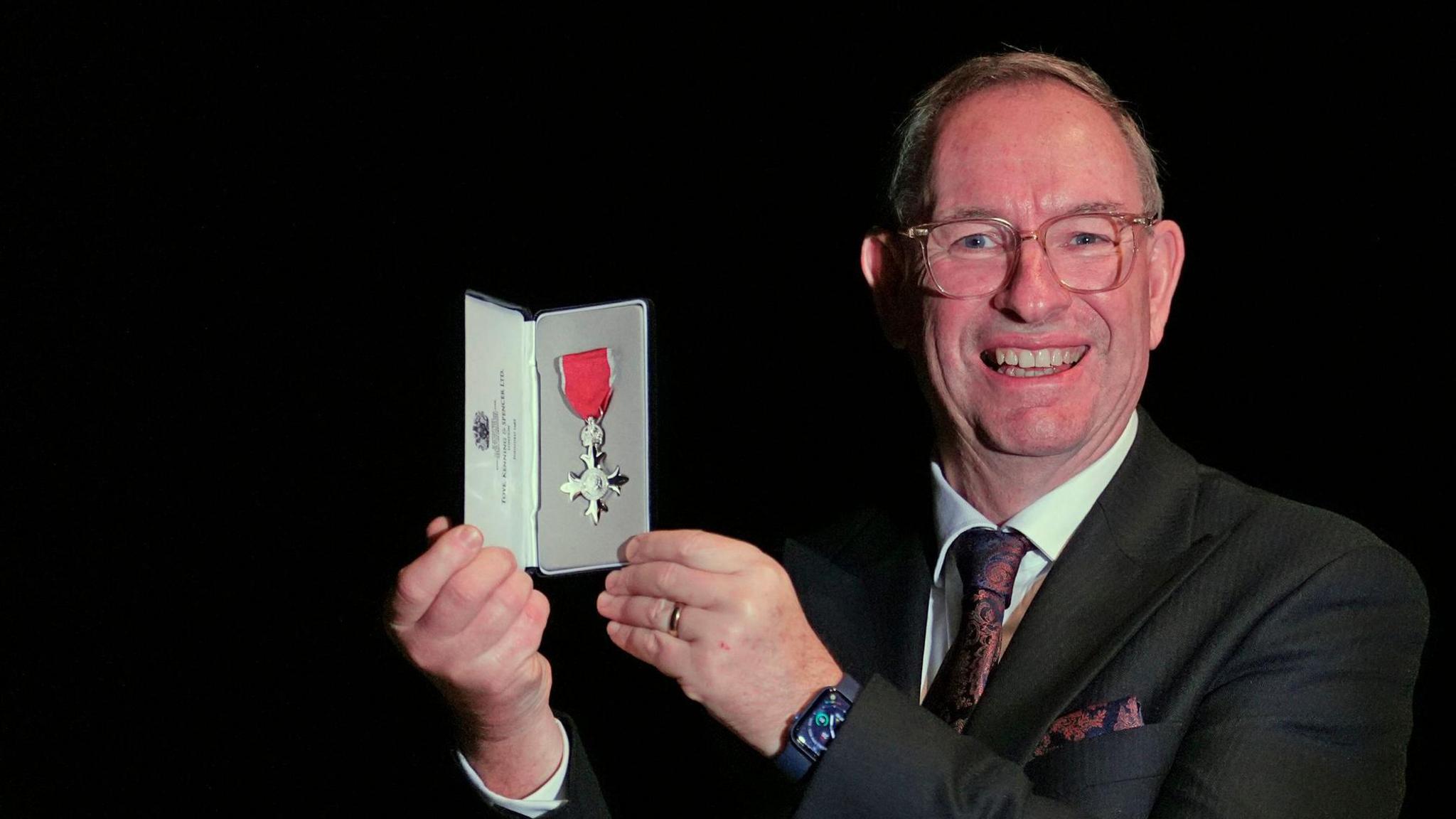 Methodist Minister, the Revd Martin Abrams, with grey hair and wearing a black suit and brown silk tie and glasses holds his Member of the Order of the British Empire (MBE) at an investiture ceremony at Windsor Castle. He is smiling.