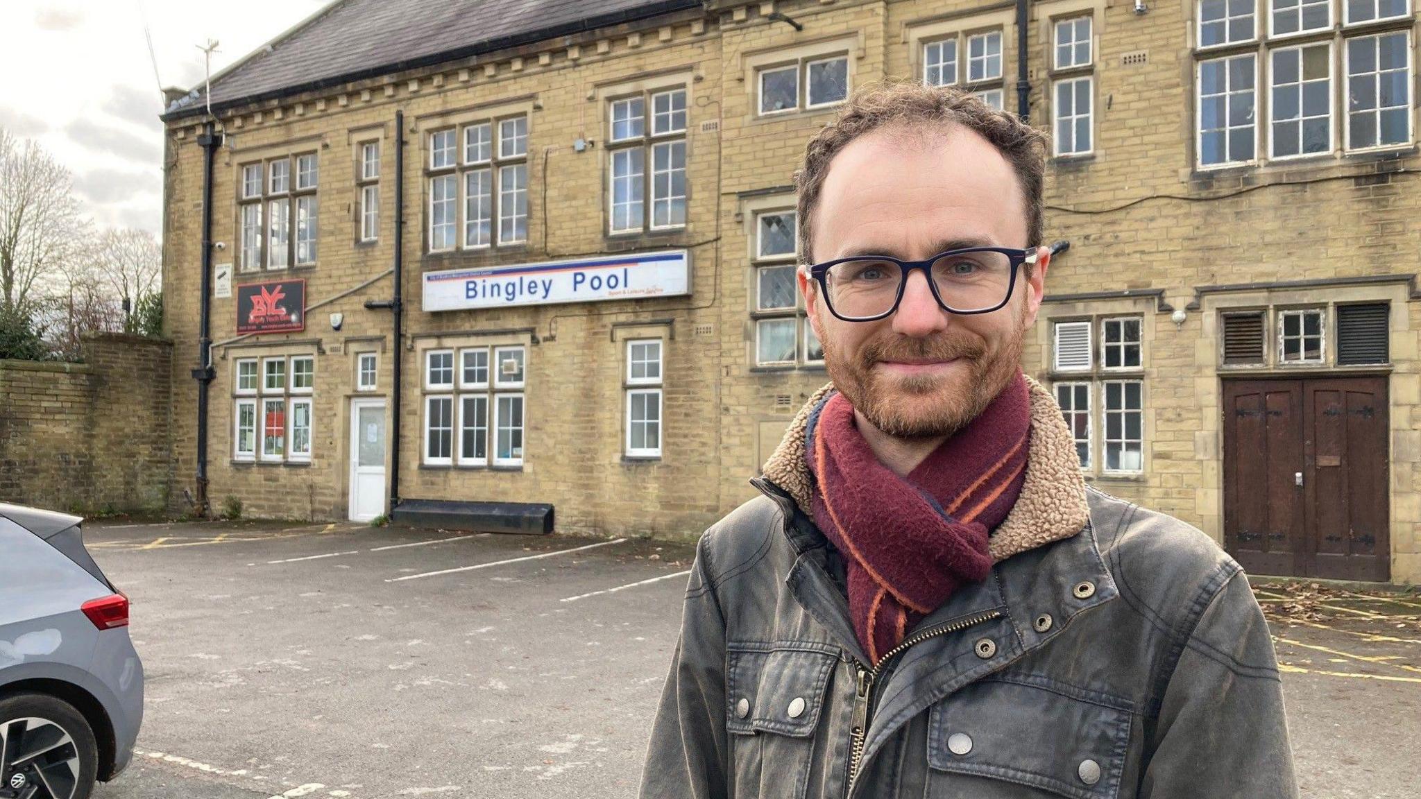 A man with brown hair and black-rimmed glasses wearing a grey jacket and purple and orange-striped scarf standing outside a faded yellow Yorkshire stone building.  