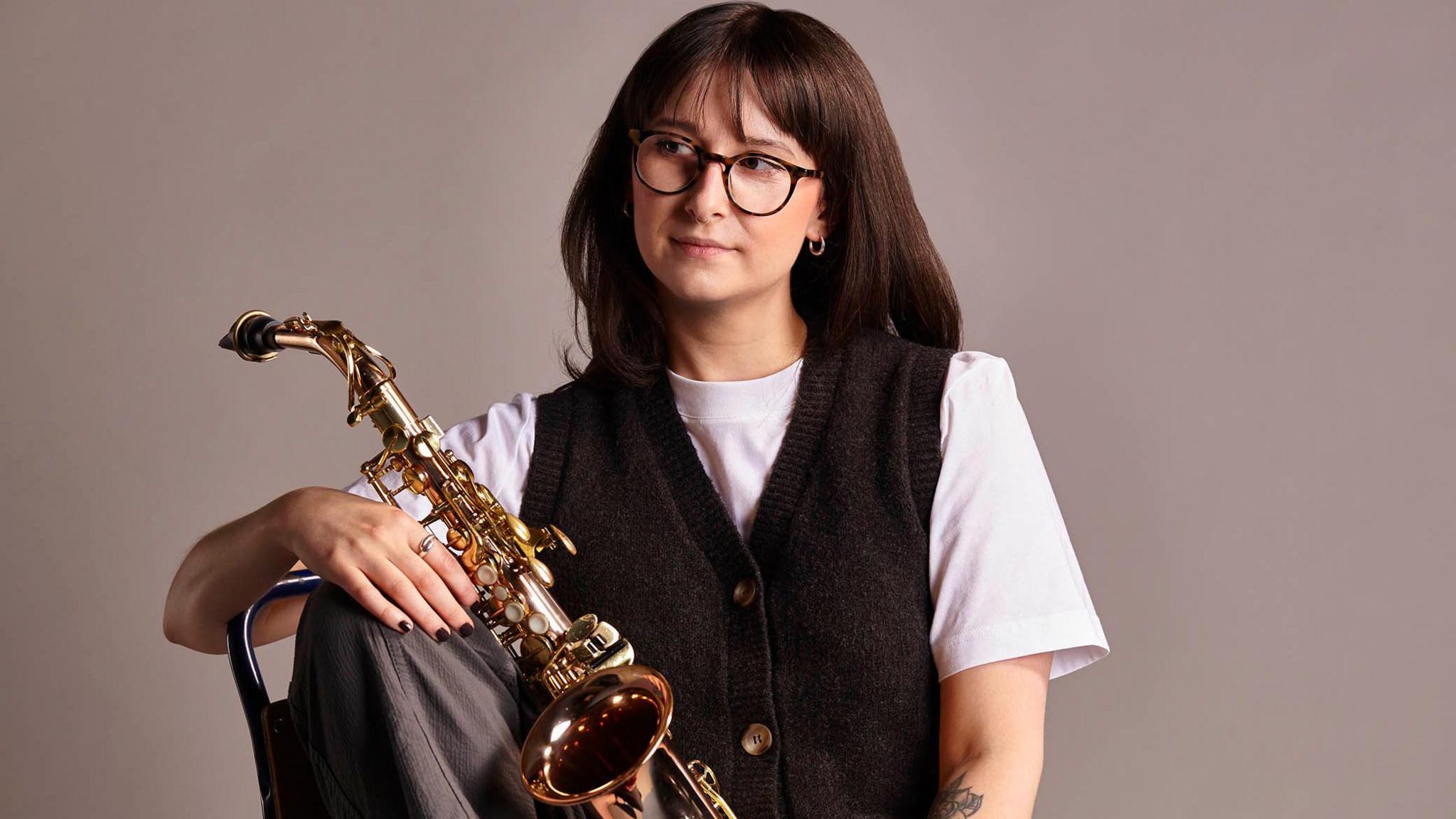 A lady with brown hair and glasses is holding a saxophone looking to the left