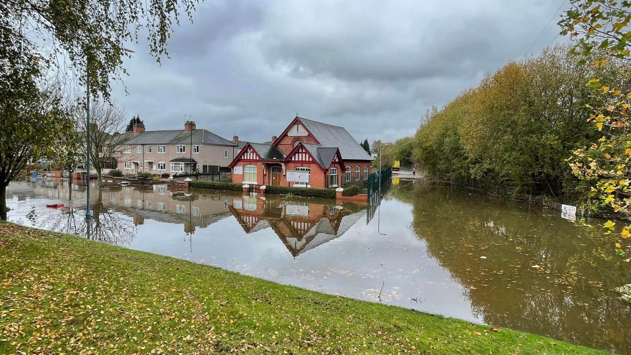 A red brick church and a few houses sit surrounded by water. Their reflections can also bee seen in the water. The water stretches across a road up to a grass verge covered in leaves. Leaves are also floating in the water. The church is on a corner with another water covered road next to it. An emergency vehicle and a figure can be seen in the distance