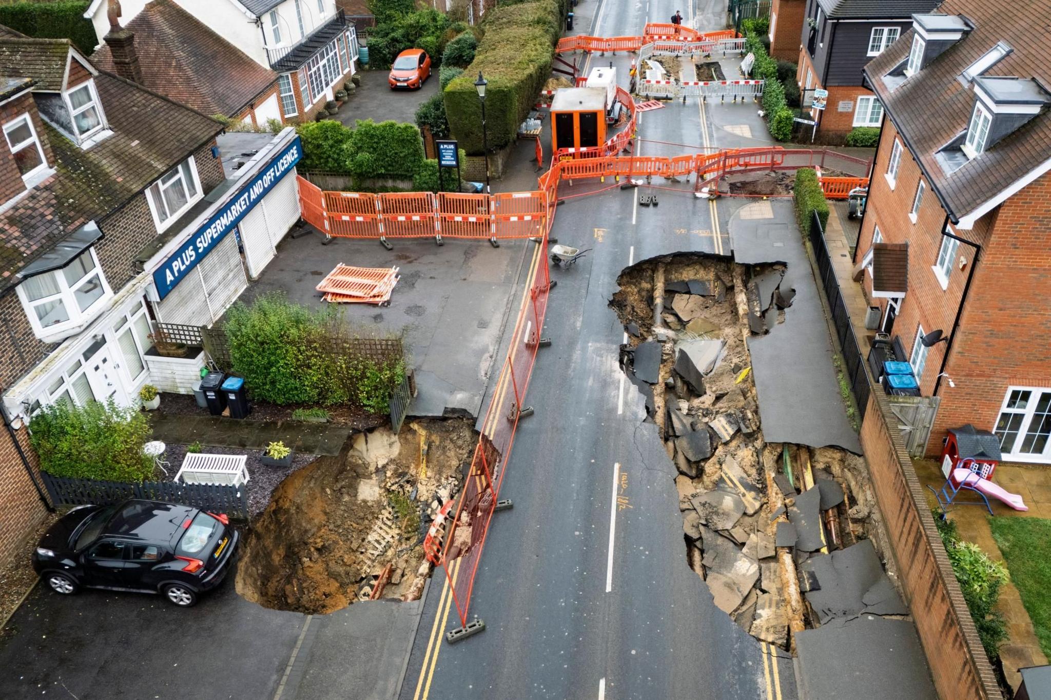 A picture taken with a drone shows sinkholes in Godstone, Surrey, UK. The largest one is at least 65ft (20m) long and is directly in front of properties on the road. A car is teetering dangerously close to the edge of another large sinkhole on the opposite side of the road. The road has been cordoned off.