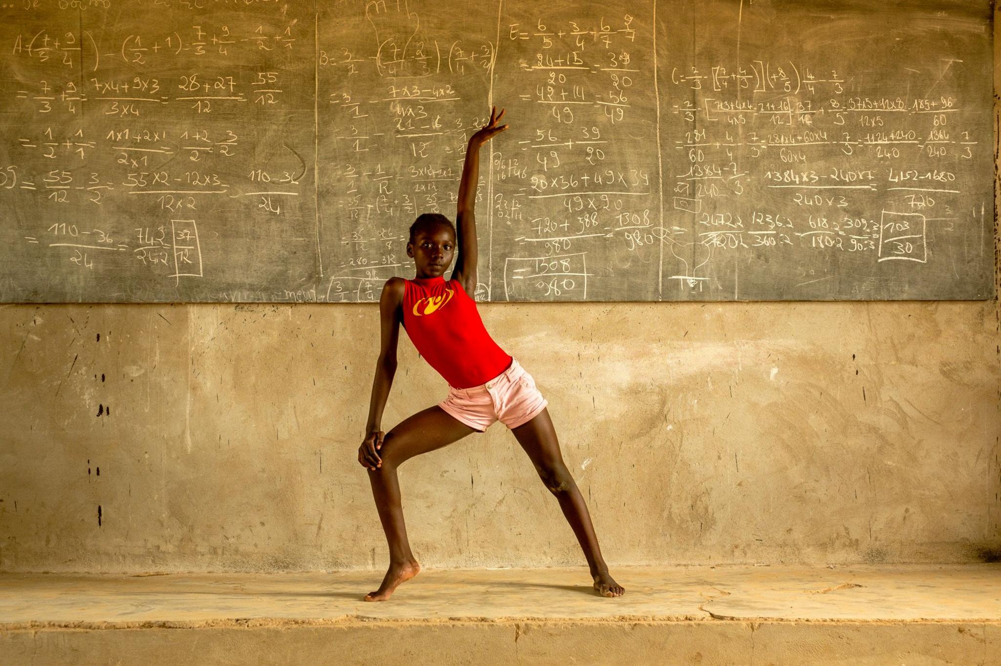 Achta practices a gymnastics pose at the school in Toukra, she poses with bare feet in front of a massive black board covered in chalk equations