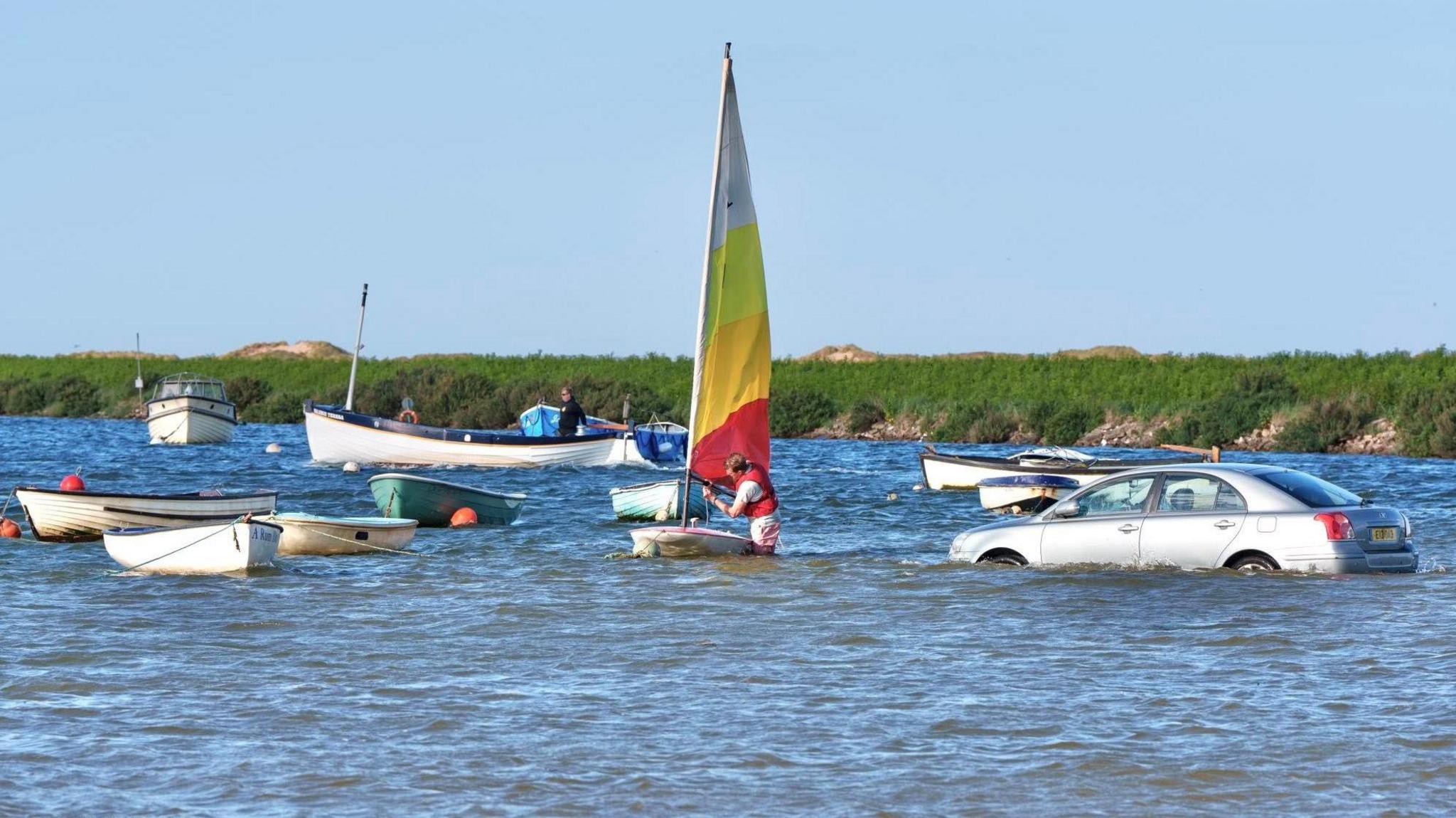 A man is standing in the water with a llife jacket on adjusting a brightly-coloured striped sail to a small boat. Boats are bobbing in the water around him and a silver saloon-type car is submerged in the water.