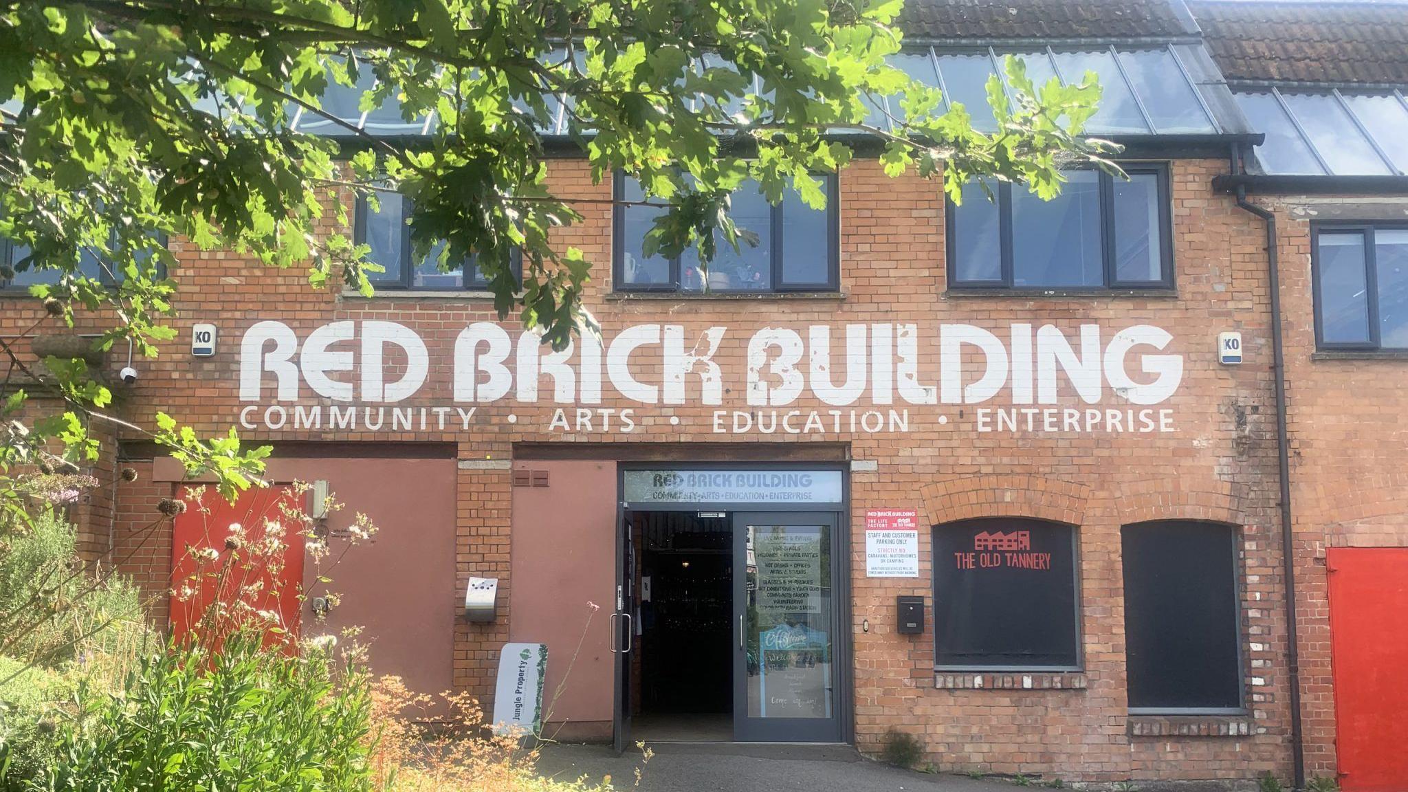 The outside of the Red Brick Building entrance with its name above the door and some foliage on the left foreground