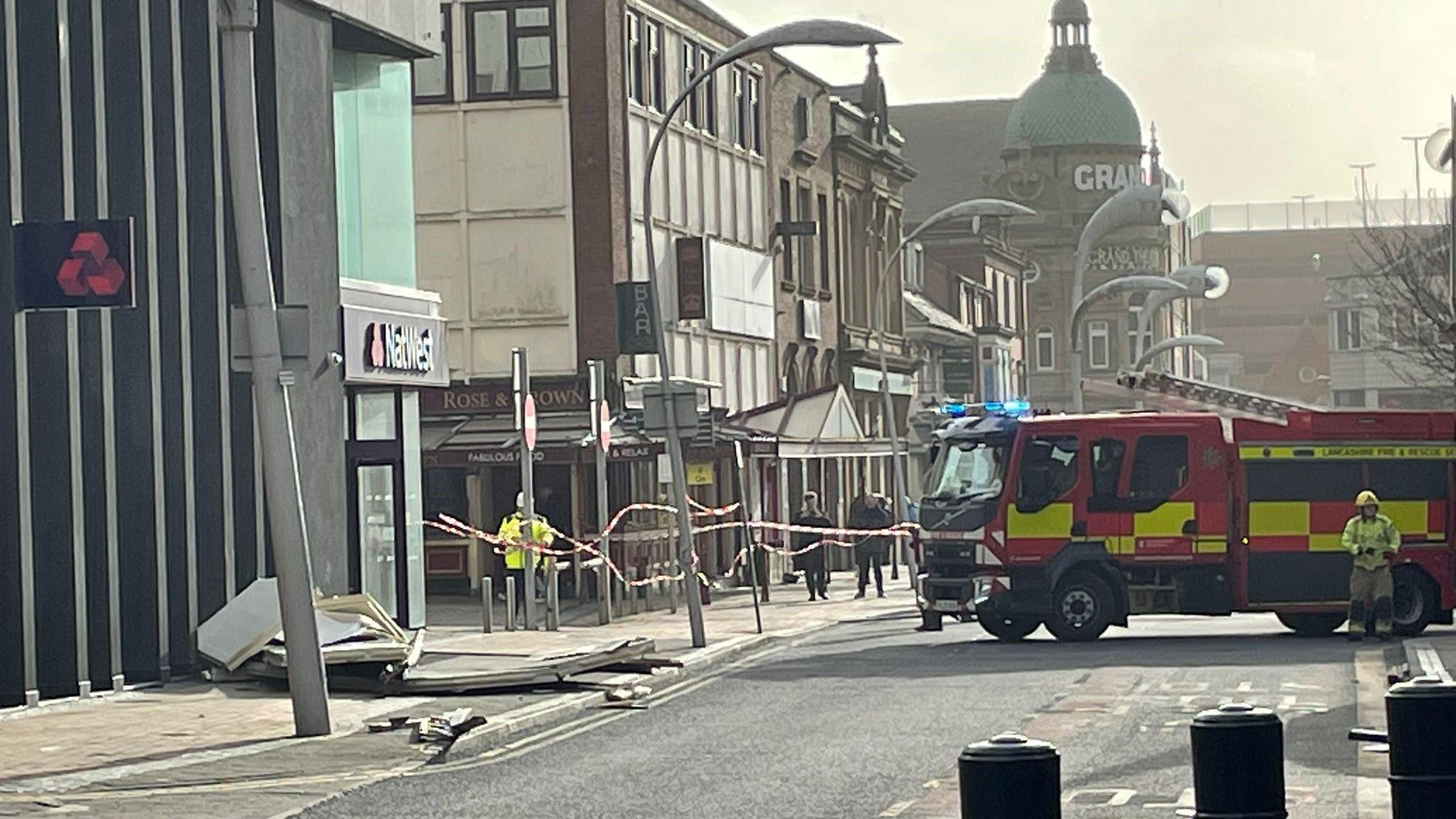 A road blocked off by emergency services. A fire engine can be seen next to a building where part of the facade has fallen off and is on the pavement next to Natwest bank.