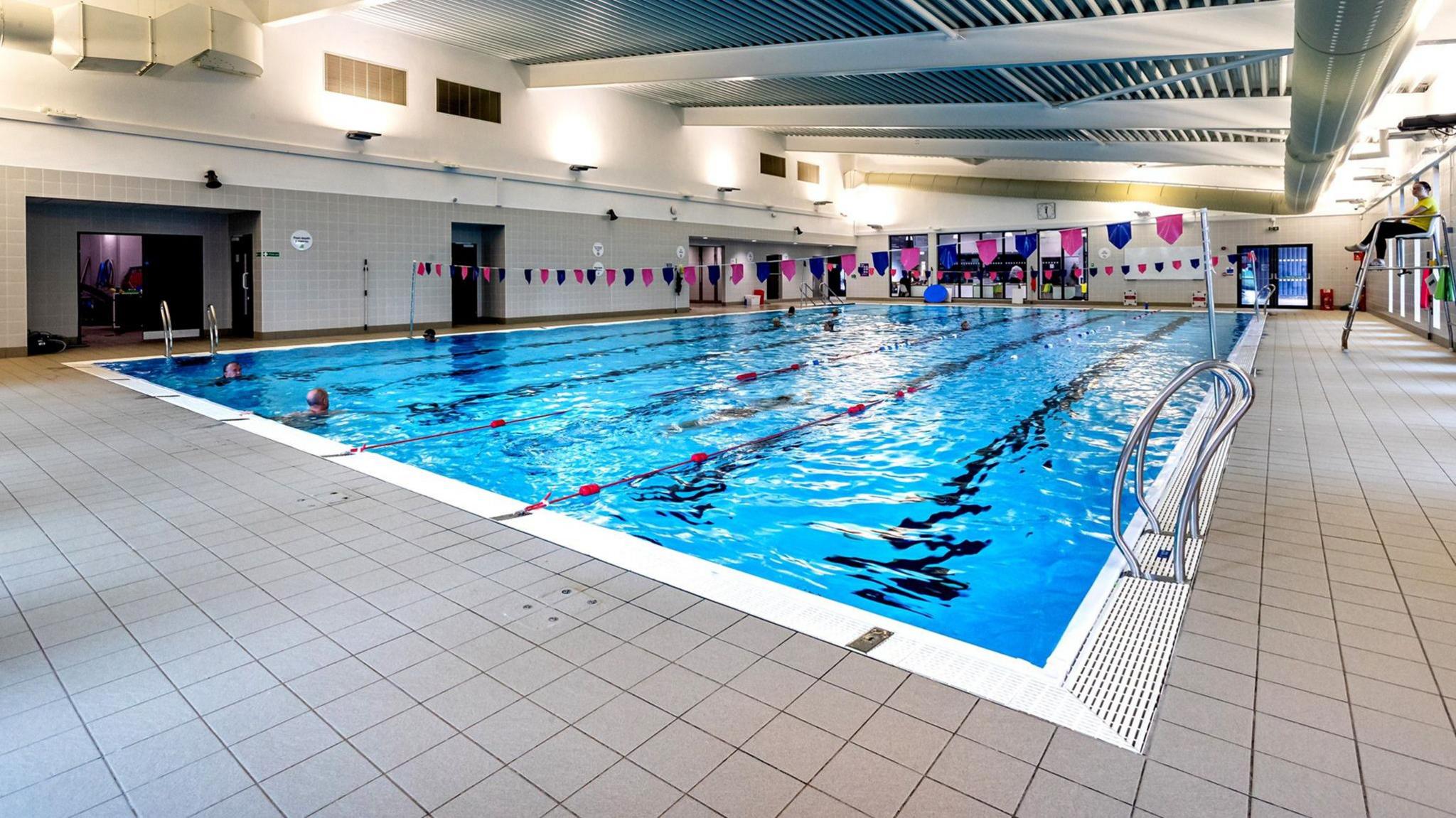 An image of Ripon's swimming pool, surrounded by beige tiling and blue, red and white bunting strung across the pool.