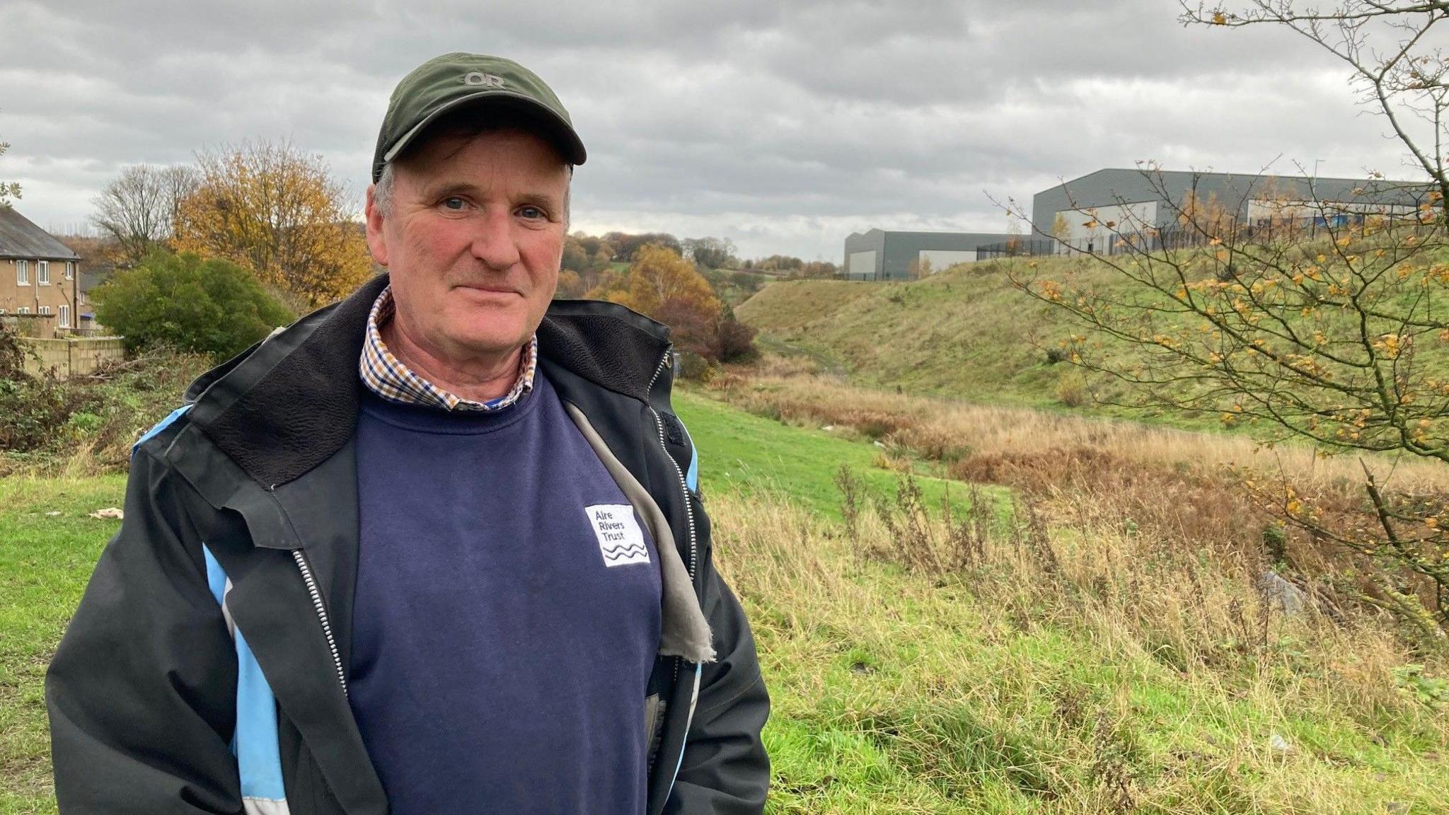 A man wearing a peaked cap and blue jumper and black coat standing in a grassy field next to a line of yellowing reeds 