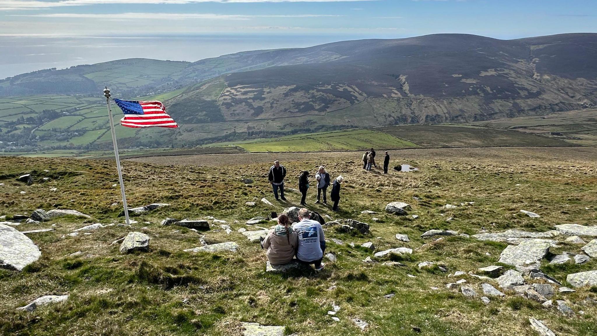 An American flag flying to the left, with a number of people sitting and standing on rocks and grass, with hills and the sea in the distance. 