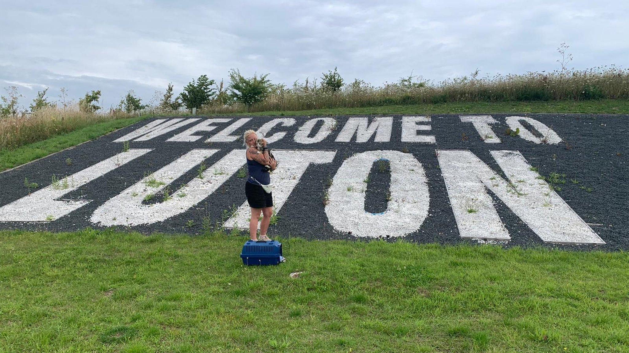 Georgie Dunt with her cat Max, standing next to a sign which says Welcome To Luton