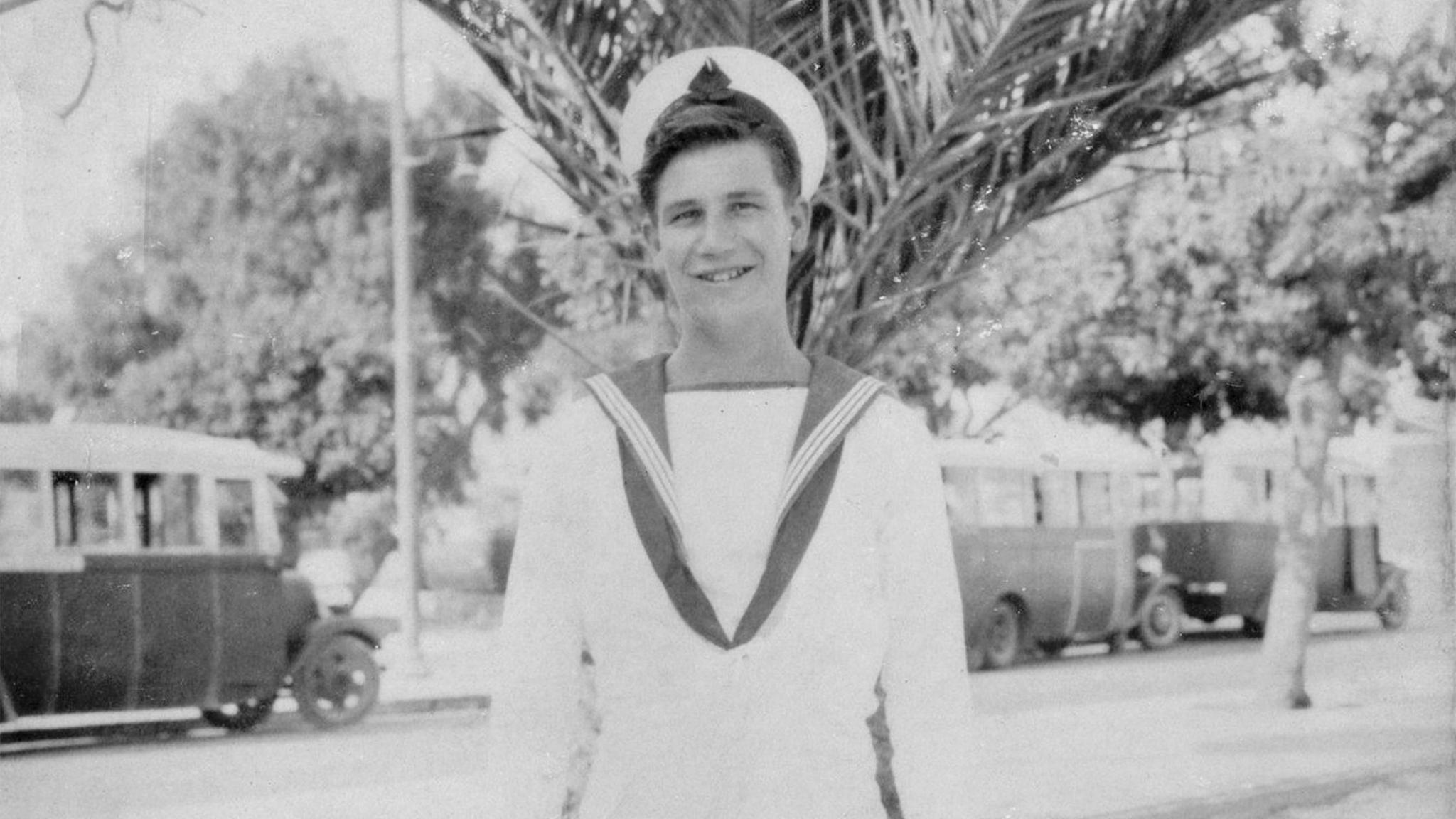 A black and white picture of George Chandler as a young man, wearing a white tropical naval ratings uniform, posing in front of a palm tree, with three buses in the background.