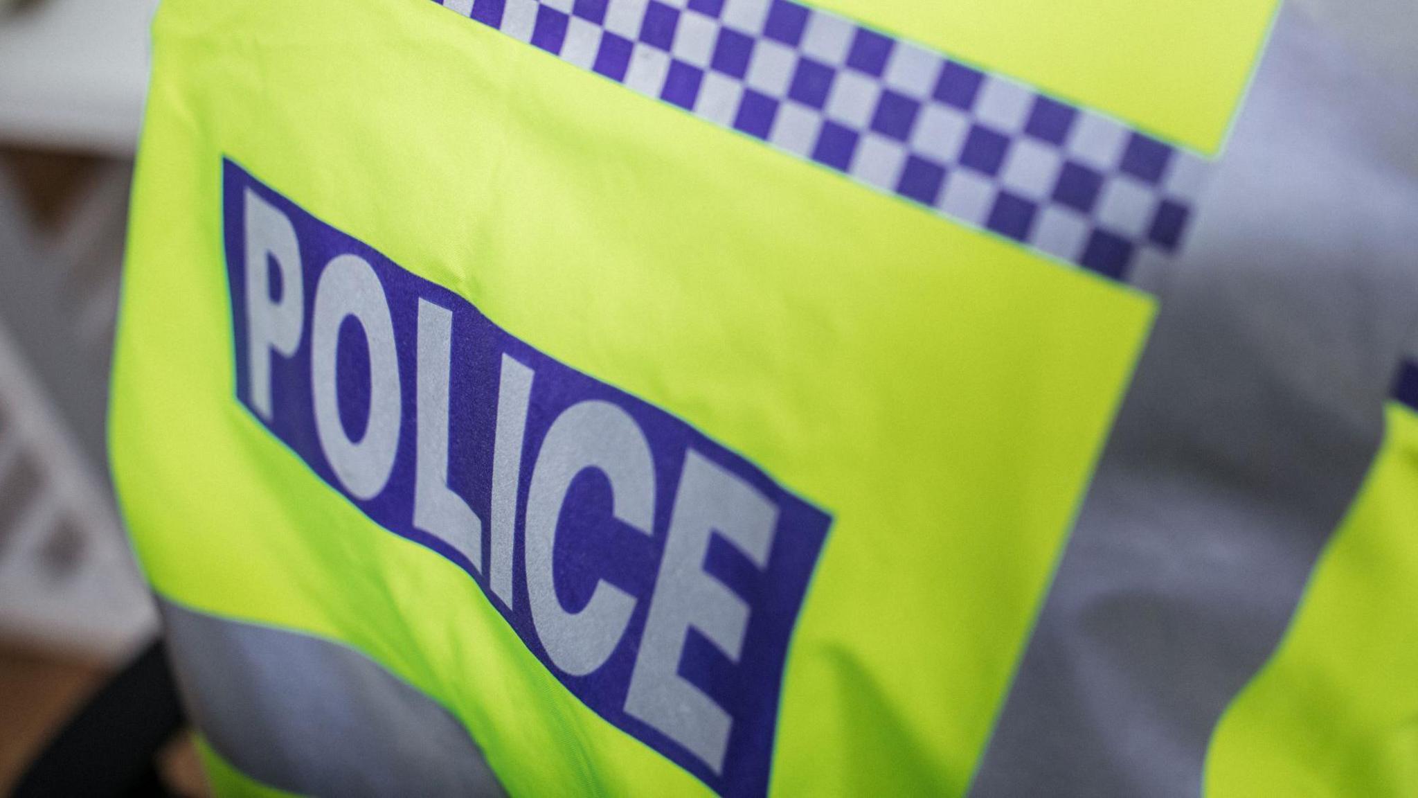 The back of a yellow hi-viz police jacket, it has the word Police in grey writing atop a blue background