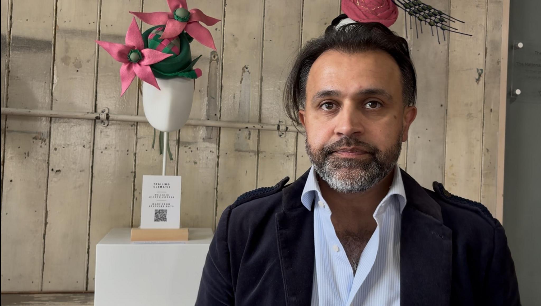Billy Hussain with medium-length brow hair, and a dark brown beard, wearing a black jacket and blue shirt. He is standing in front of two hats on display at the Hat Factory, with a wooden wall behind.