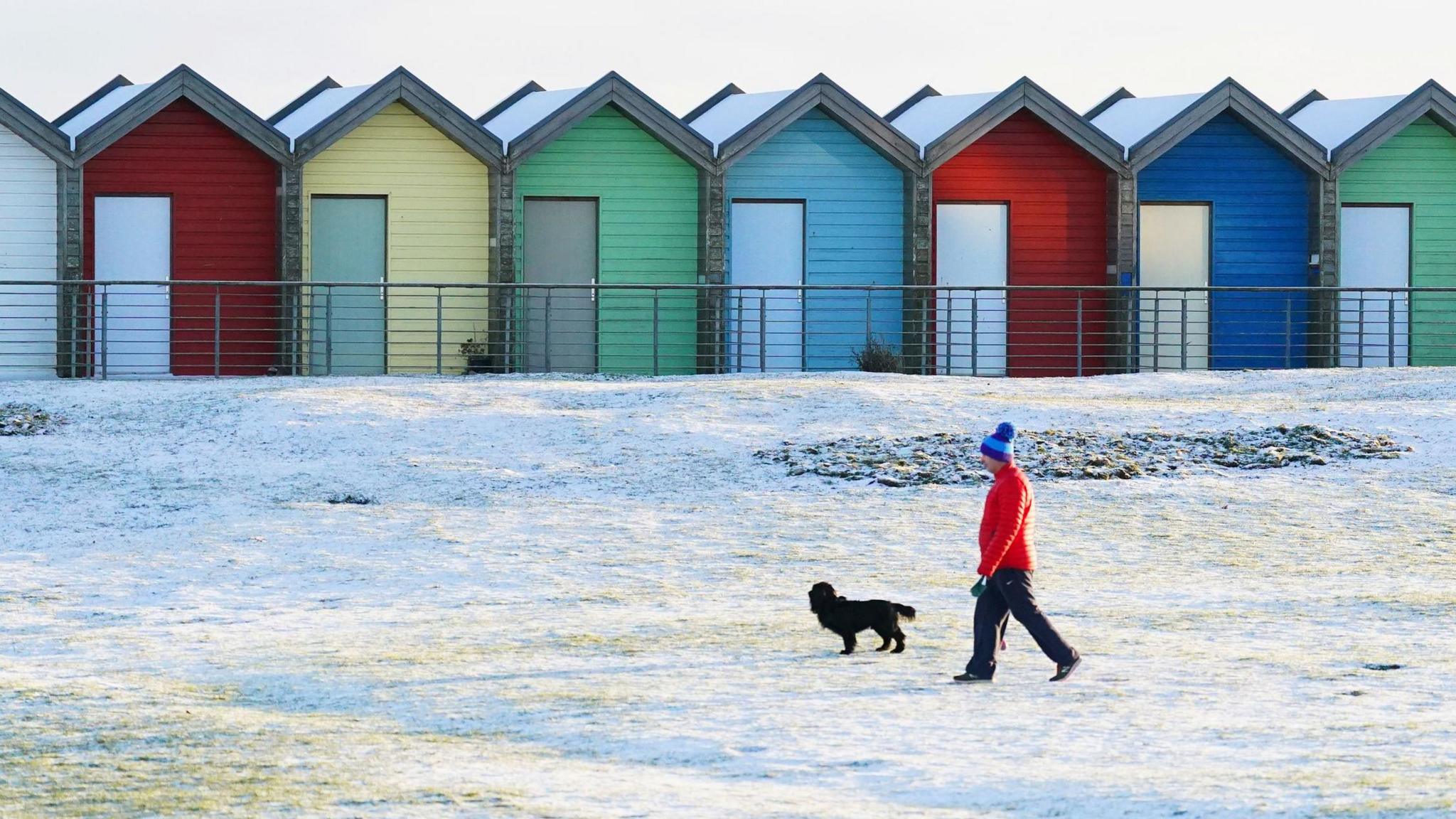 A man dressed in a red puffer jacket and blue bobble hat walks his black cocker spaniel on a snow covered beach in Blyth. He is walking past a row of brightly coloured beach huts.