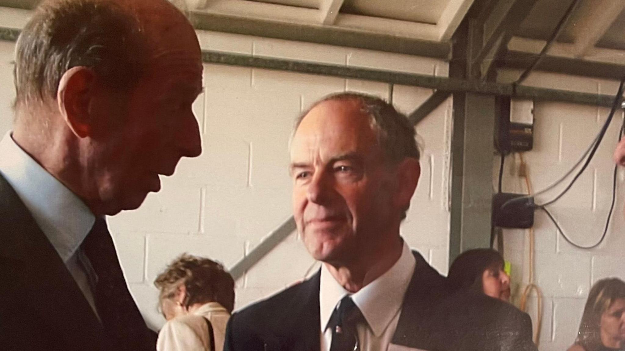 The Duke of Kent, wearing a dark suit jacket, blue shirt and tie, speaks to Mr Huggins, wearing a dark suit jacket, white shirt and dark tie, in the whitewashed lifeboat shed. Three women are in the background.