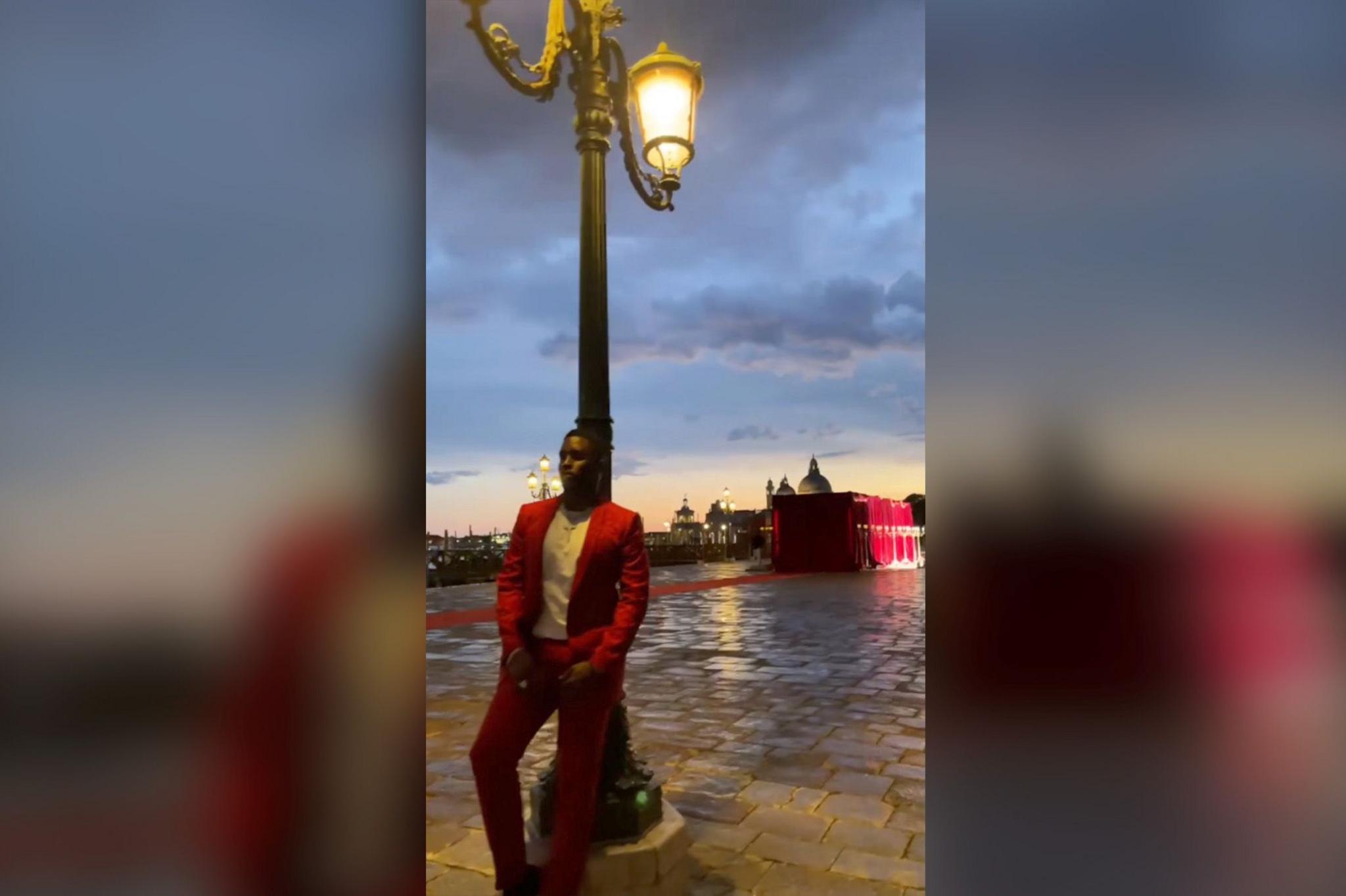 Combs dressed in a red suit and white shirt leaning against a street lamp post on a cobblestone path, with buildings and a sunset in the background.