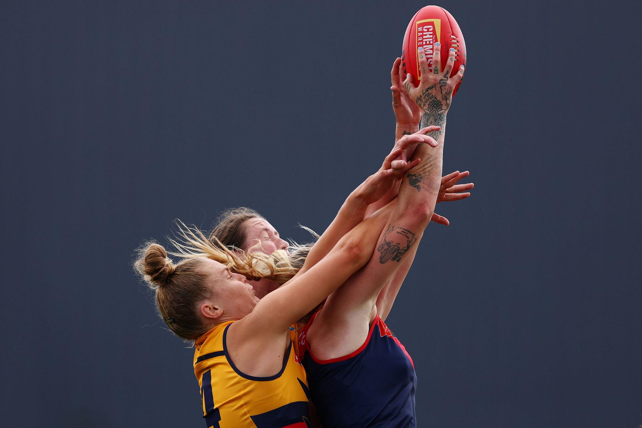 Two players competing for an Australian rules football, both reaching high with extended arms, one holding the red ball, against a plain dark background.