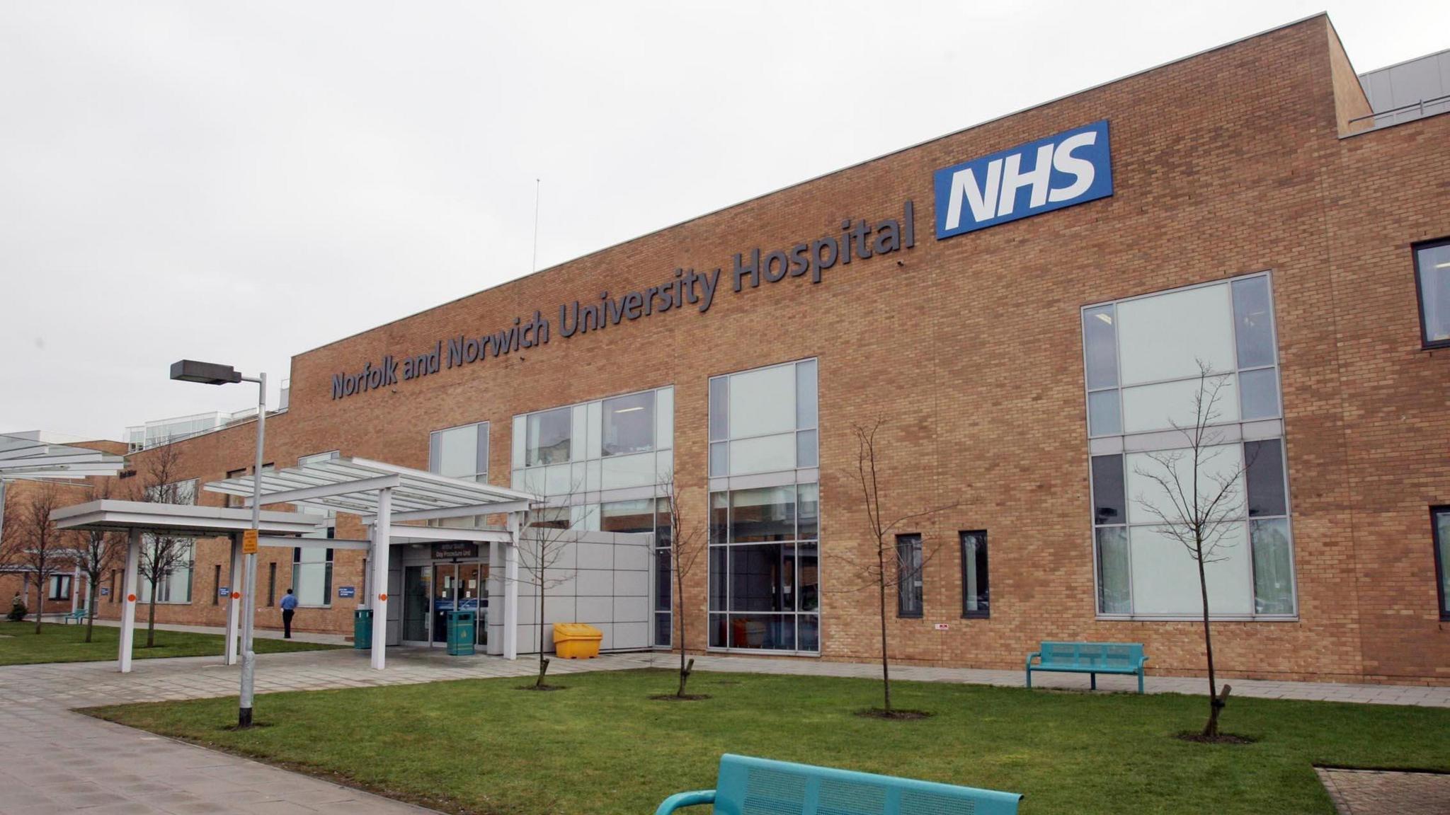 The outside of the Norfolk and Norwich University Hospital. At the top of the red-brick building a sign says Norfolk and Norwich University Hospital NHS. The building has large glass windows and a small grass area to its front. On the grass are green benches. 