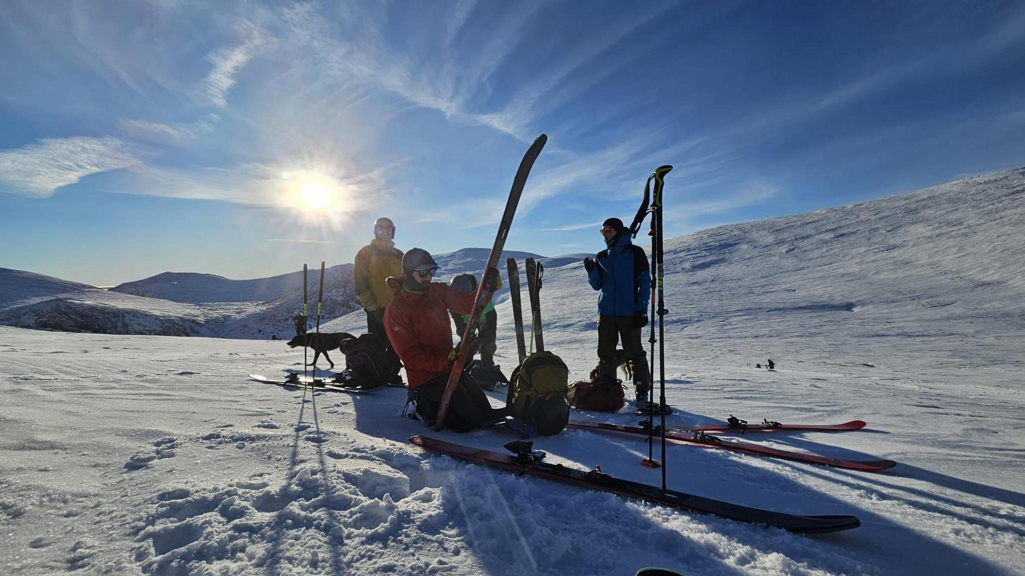 Three people working on their skis in the snow. the sun is shining in a blue sky. the group cast long shadows across the whiteness.