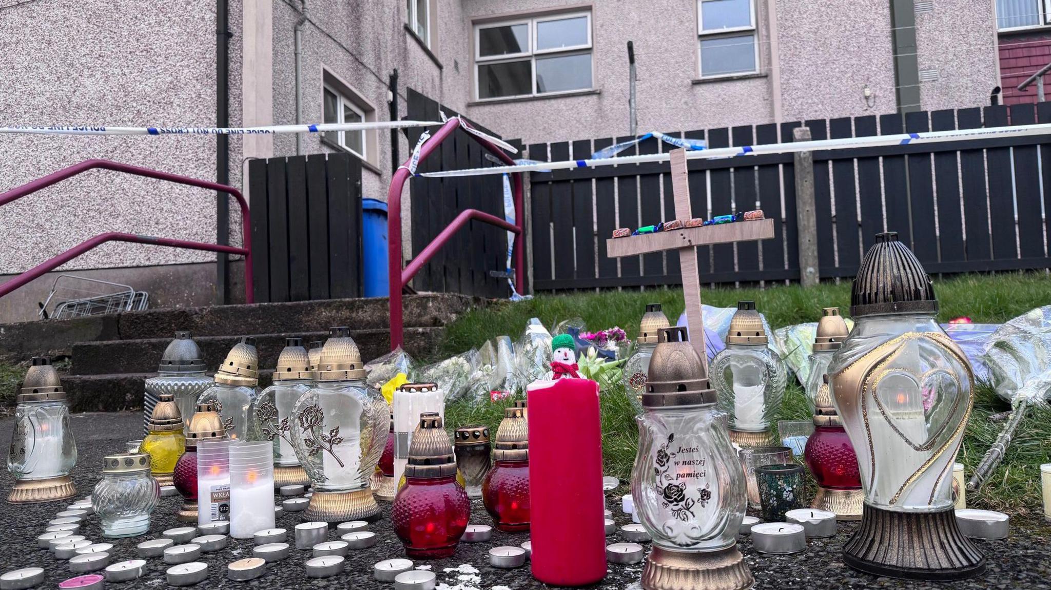 A collection of glass lanterns, candles, tealights and a small wooden cross which were left near the block of flats where a man died after an assault in Dungannon.
The flats are in the background and a white police cordon surrounds the building
