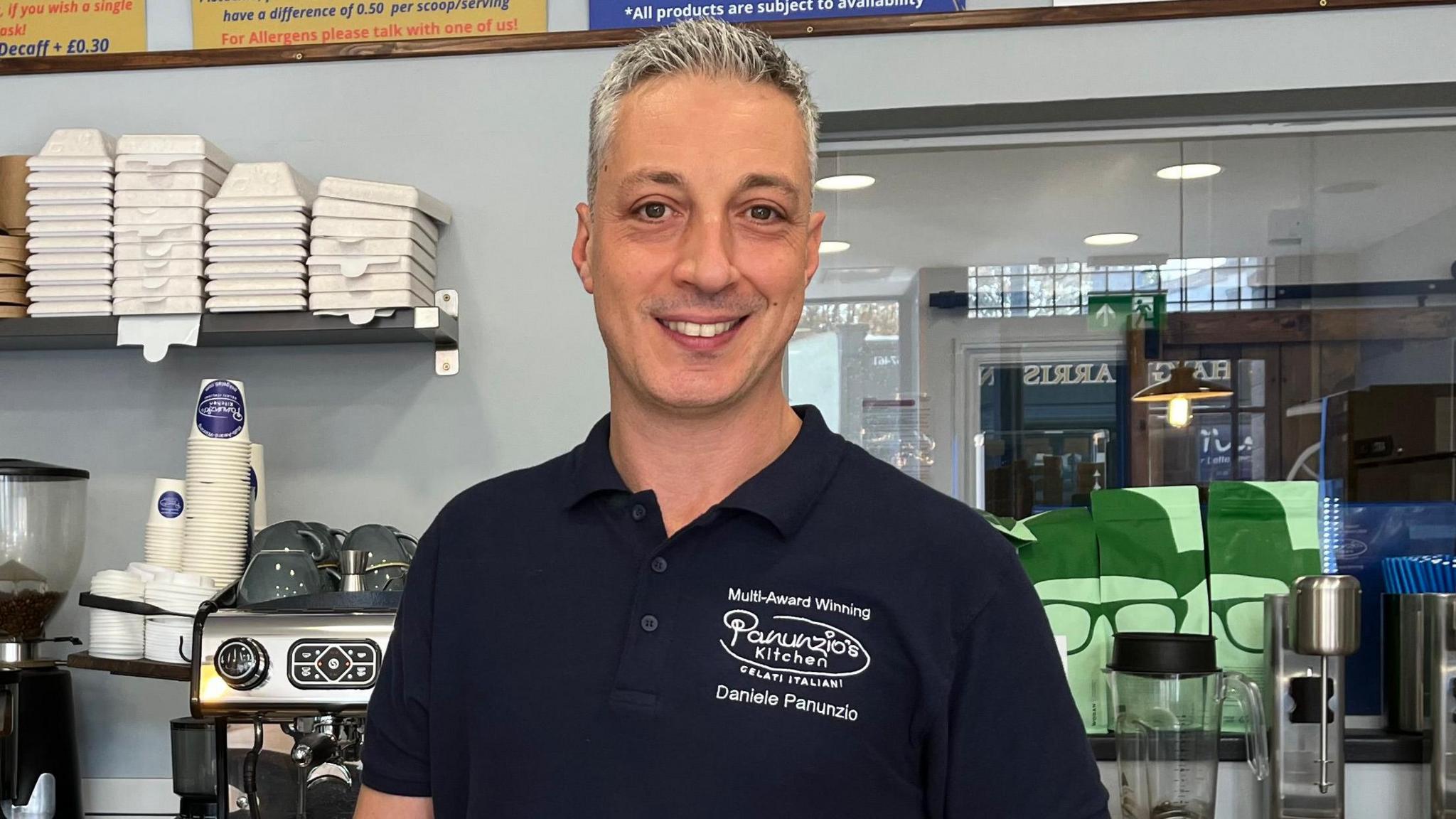 Daniele Panunzio wearing a black Panunzio's Kitchen branded polo shirt and smiling at the camera. He is standing behind the counter of his ice cream shop in Bristol, which is painted a pale blue and has shelves stacked with boxes, cups and coffee. In the foreground there is a glass jar full of baked treats and Mr Panunzio is holding a metal scoop of a pink gelato and posing for a picture.