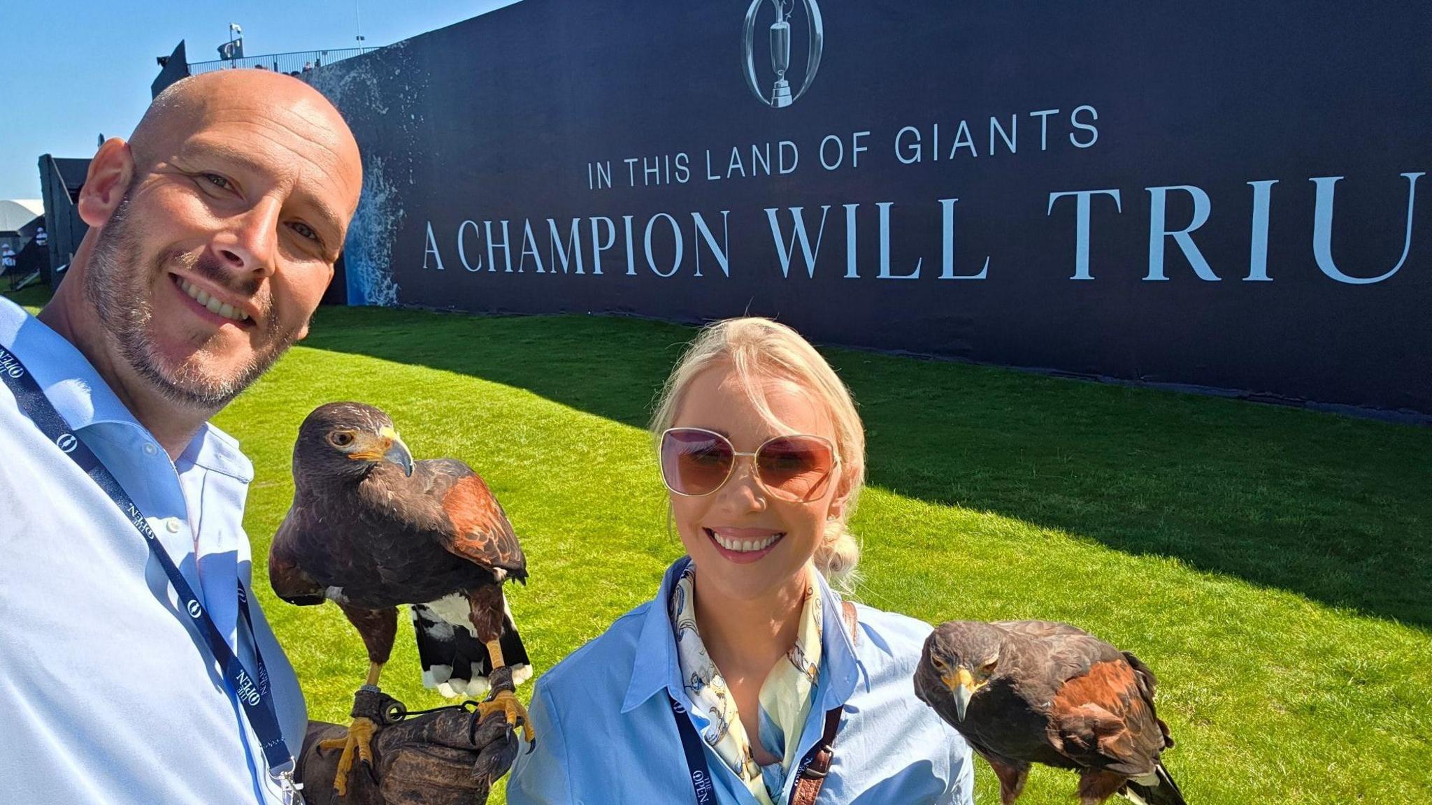 A man and woman stand side by side, each dressed in a blue shirt and each holding a Harris hawk on their well protected arms. Both people are smiling. Both birds, which are a mix of shades of brown in colour, are motionless. A sign behind them showcases the Open championship golf tournament.