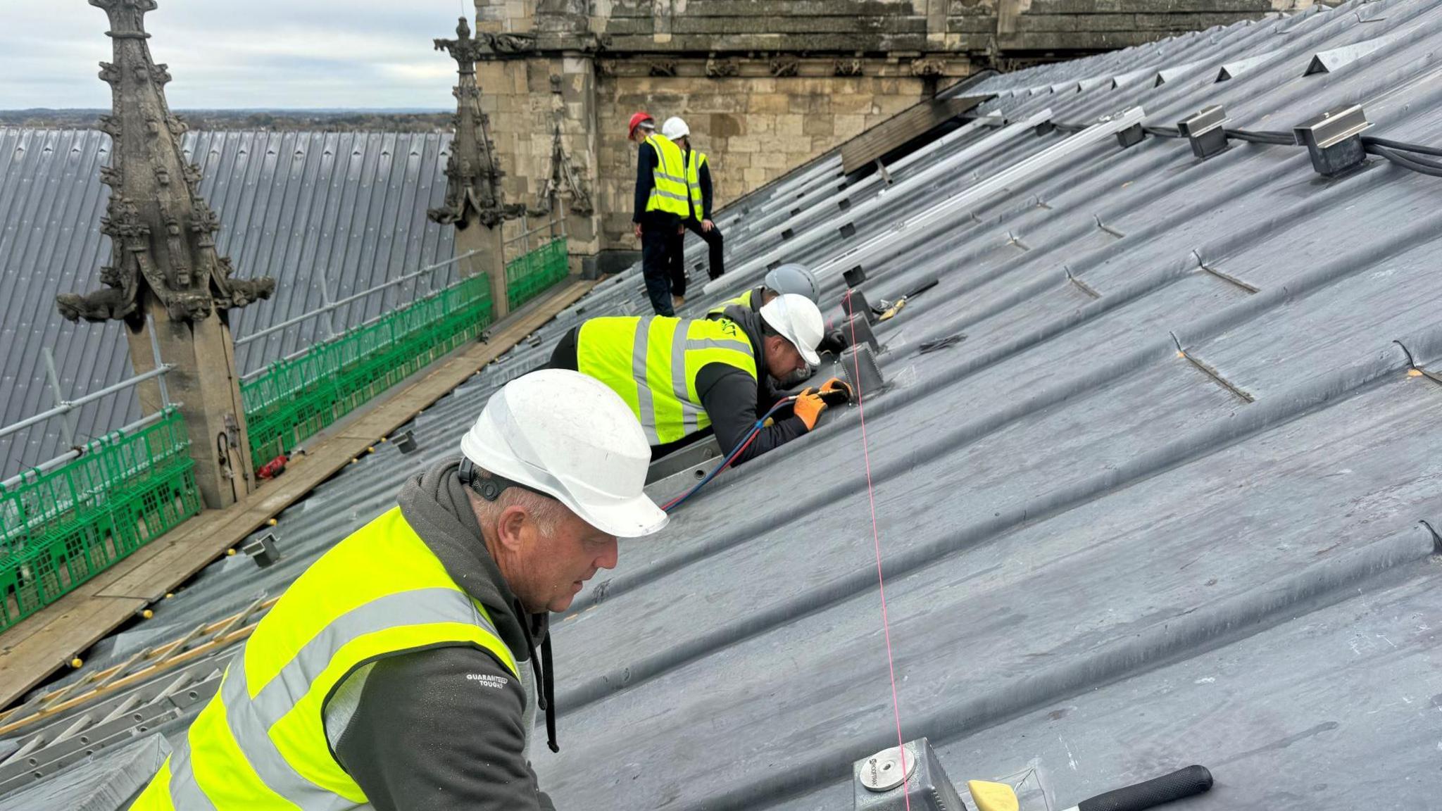 Five workers in helmets and hi-vis jackets crouched on the slanted roof of York Minster