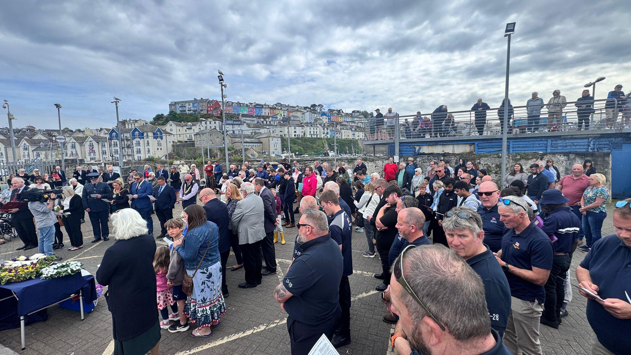 Brixham Fish Market is packed with people during the service and they have their heads bowed and are holding paper programmes. There are floral tributes on a table.