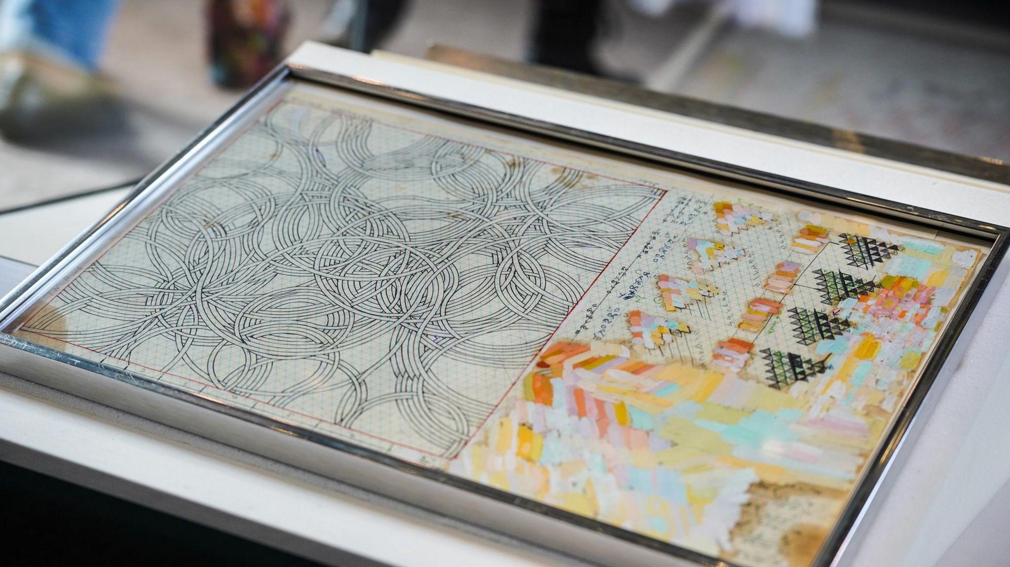 A drawing in a glass case. On the left there is lots of curved lines intersecting in black ad white. On the right there looks to be lots of pastel colours being tested, with swatches covering much of the side.