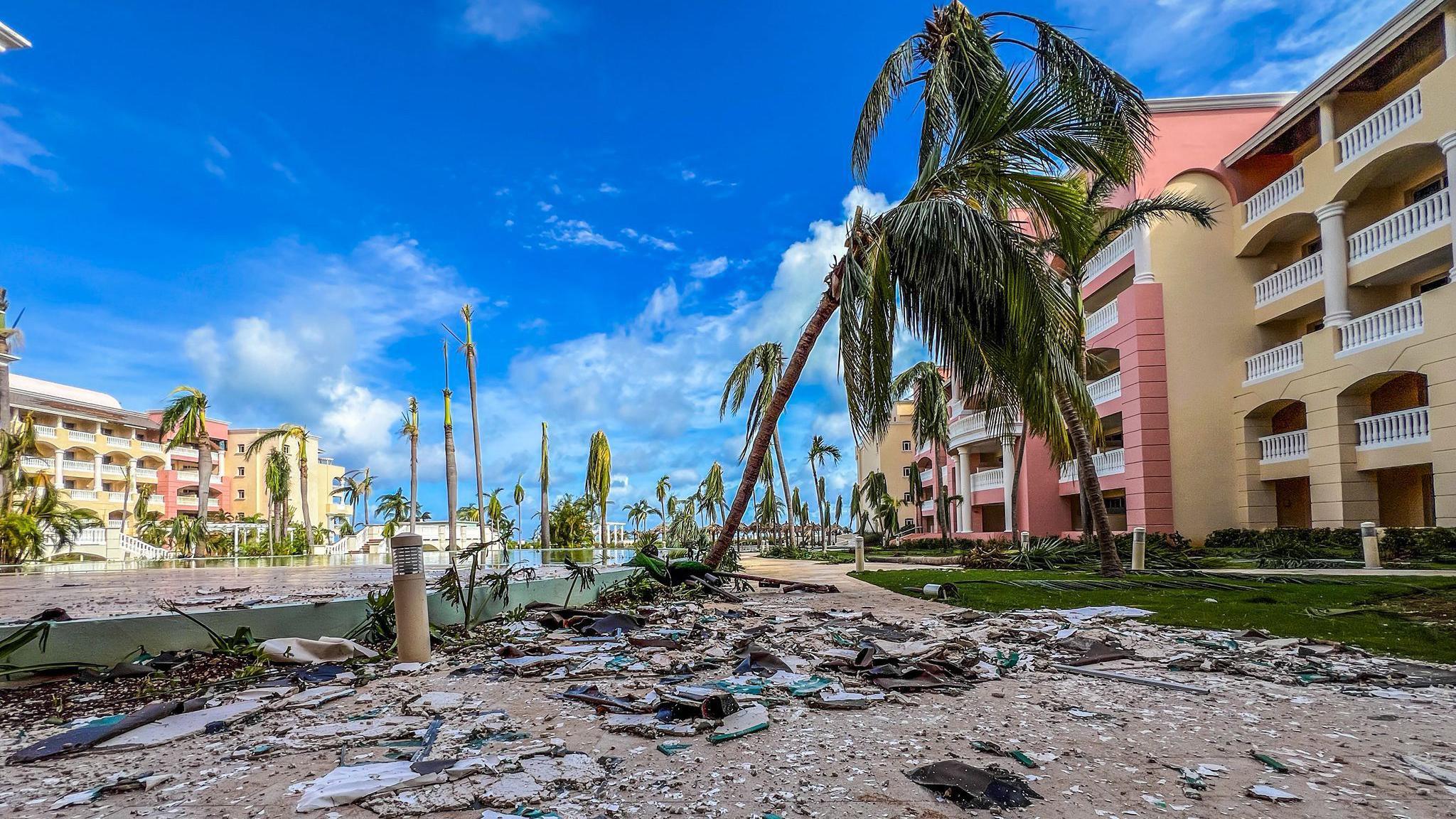 A holiday resort with modern pink and yellow buildings is covered in litter. Palm trees are ripped up on the ground or are leaning over ready to fall