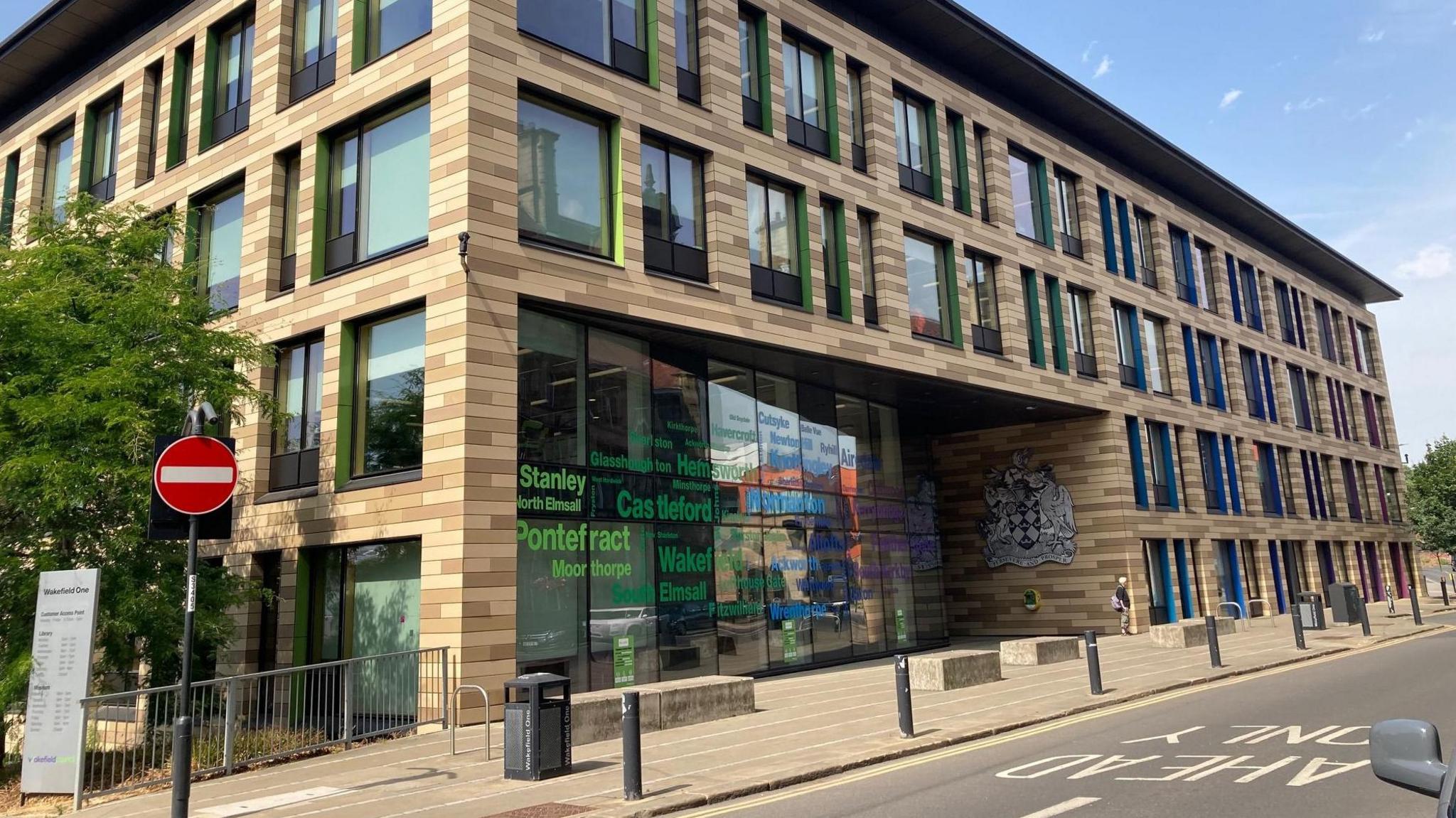 The head office of Wakefield Council, it is brown building with an exposed brick finish. A glass wall can be seen with the names of areas the council covers.