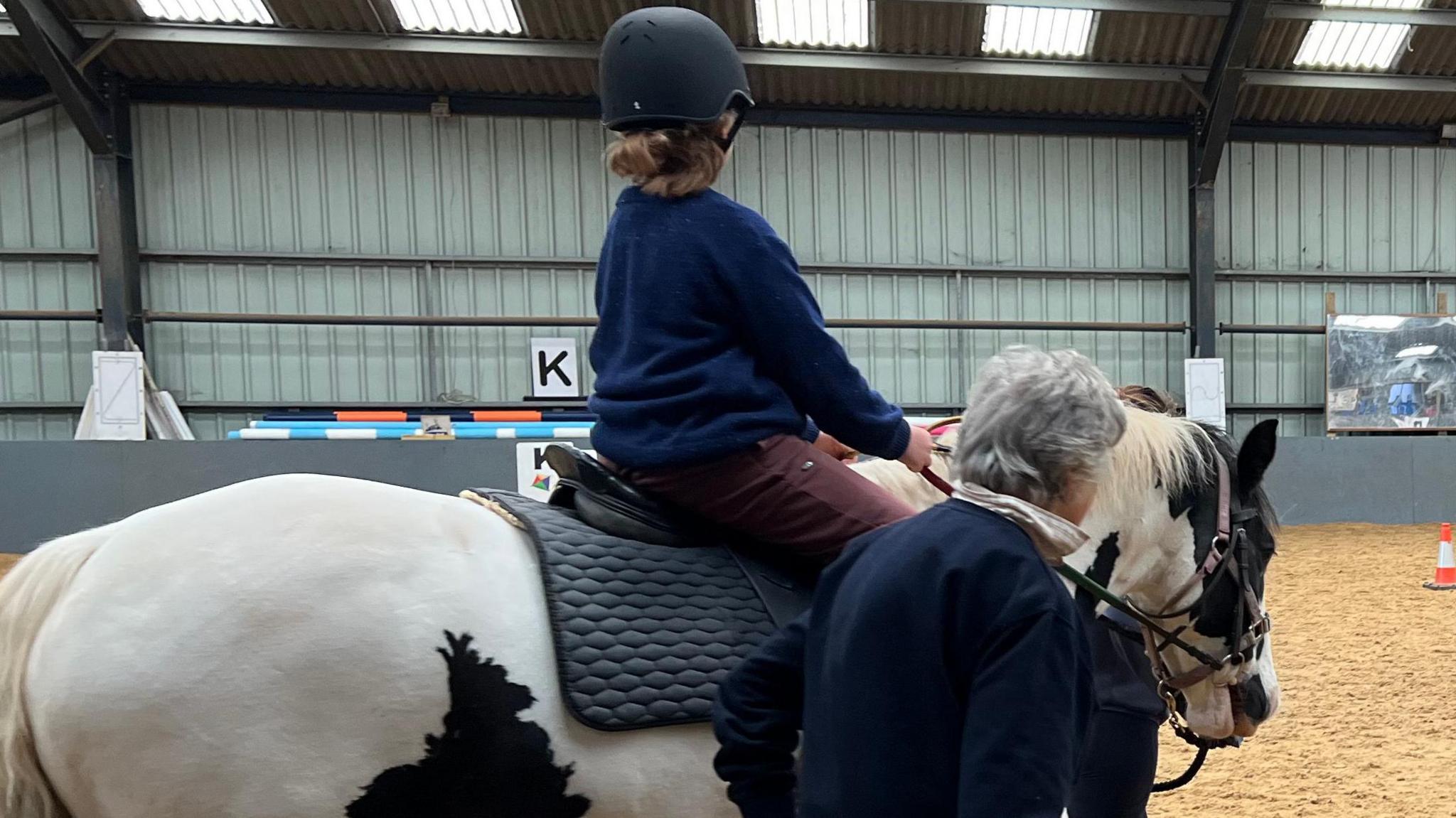 A view of the horse riding school. There are two horses, with two riders on them.  One is on the left, and is black and white, and the other is all black. The ground is light brown and there are blue and white tubes on the ground. There are people stood either side of the horses, leading them around the school.