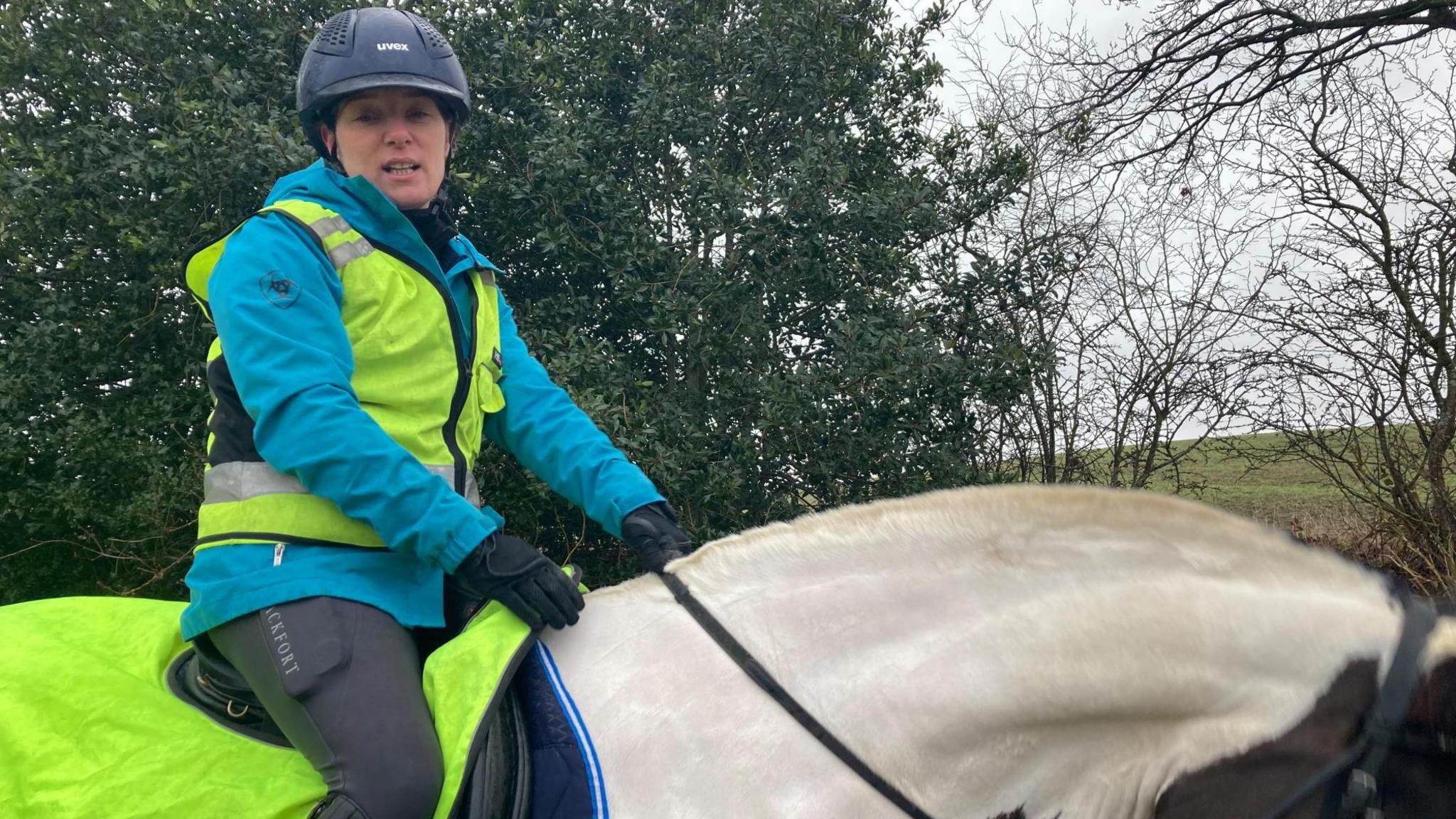 A woman in a black helmet and fluorescent jacket sits on a white horse looking down at the camera