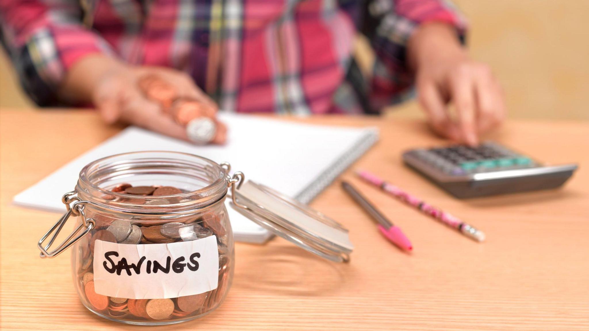 Jar of coins with sticker saying "savings" in the foreground, with a hand, coins, calculator, pen, pencil and paperwork behind.