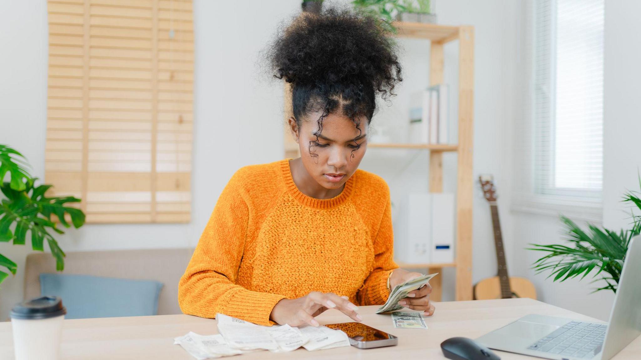 A young woman with dark hair tied up wearing an orange jumper holding cash with her phone, bills and laptop around her.