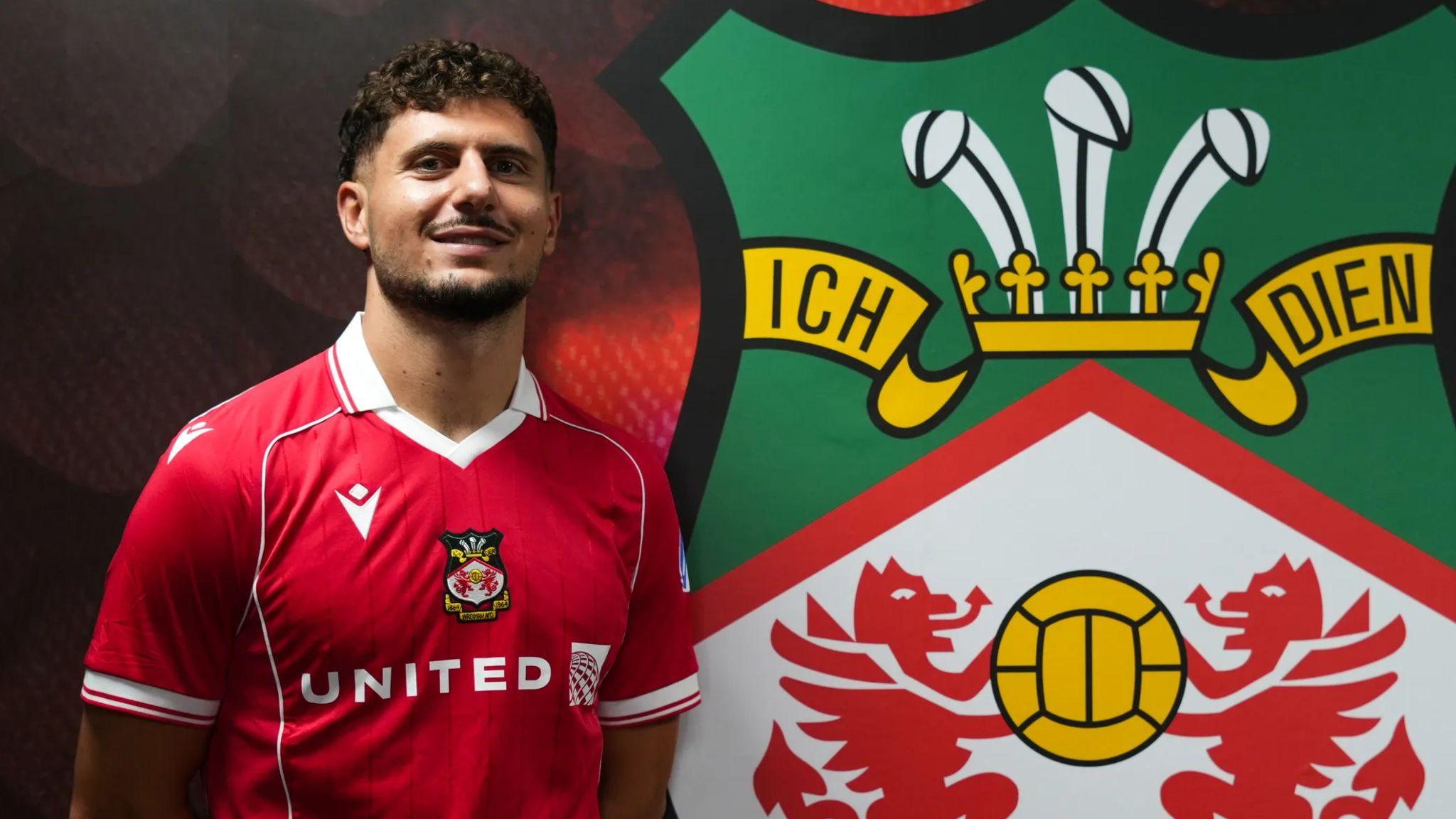Liberato Cacace poses in a Wrexham shirt alongside the club logo