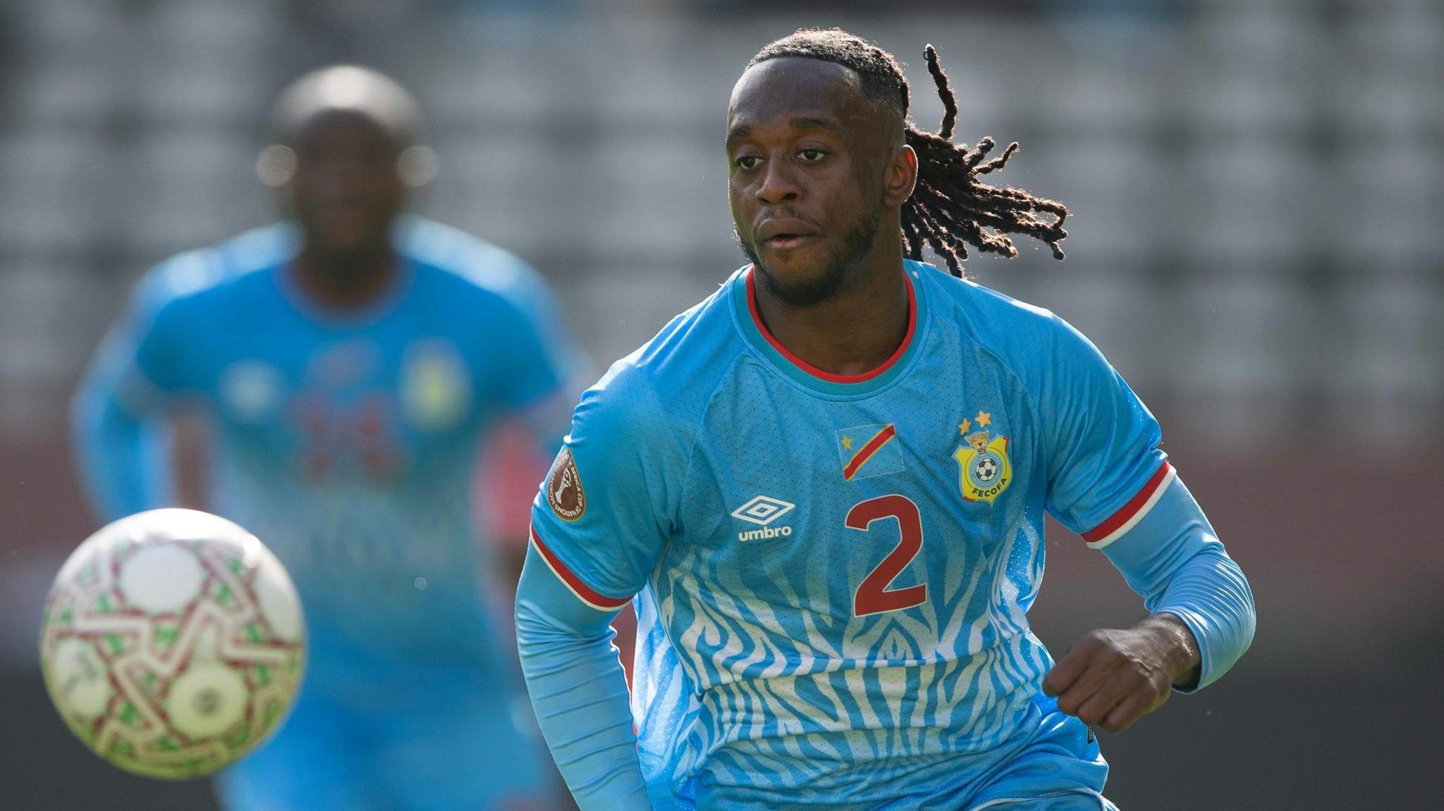 Aaron-Wan Bissaka, wearing a light blue DR Congo shirt with a red number two on the chest, is seen running as he chases a ball that is out of focus in the foreground. A DR Congo team-mate is seen in the background out of focus