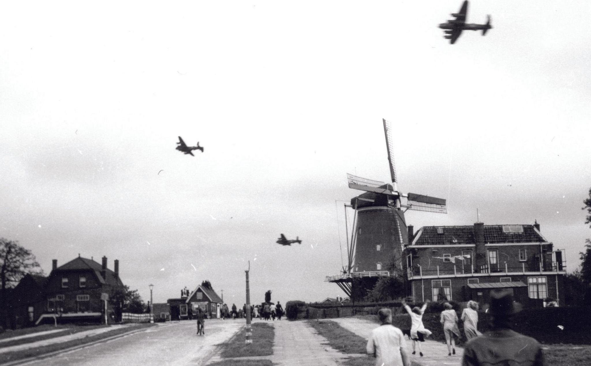 A black and white photograph of three planes flying overhead in the sky. There is a road on the left and two paths on the right where a group of three girls are walking, one with her hands in the air waving to the plane. There are groups of people in the distance. There is a house on the left and a building and a windmill on the right.