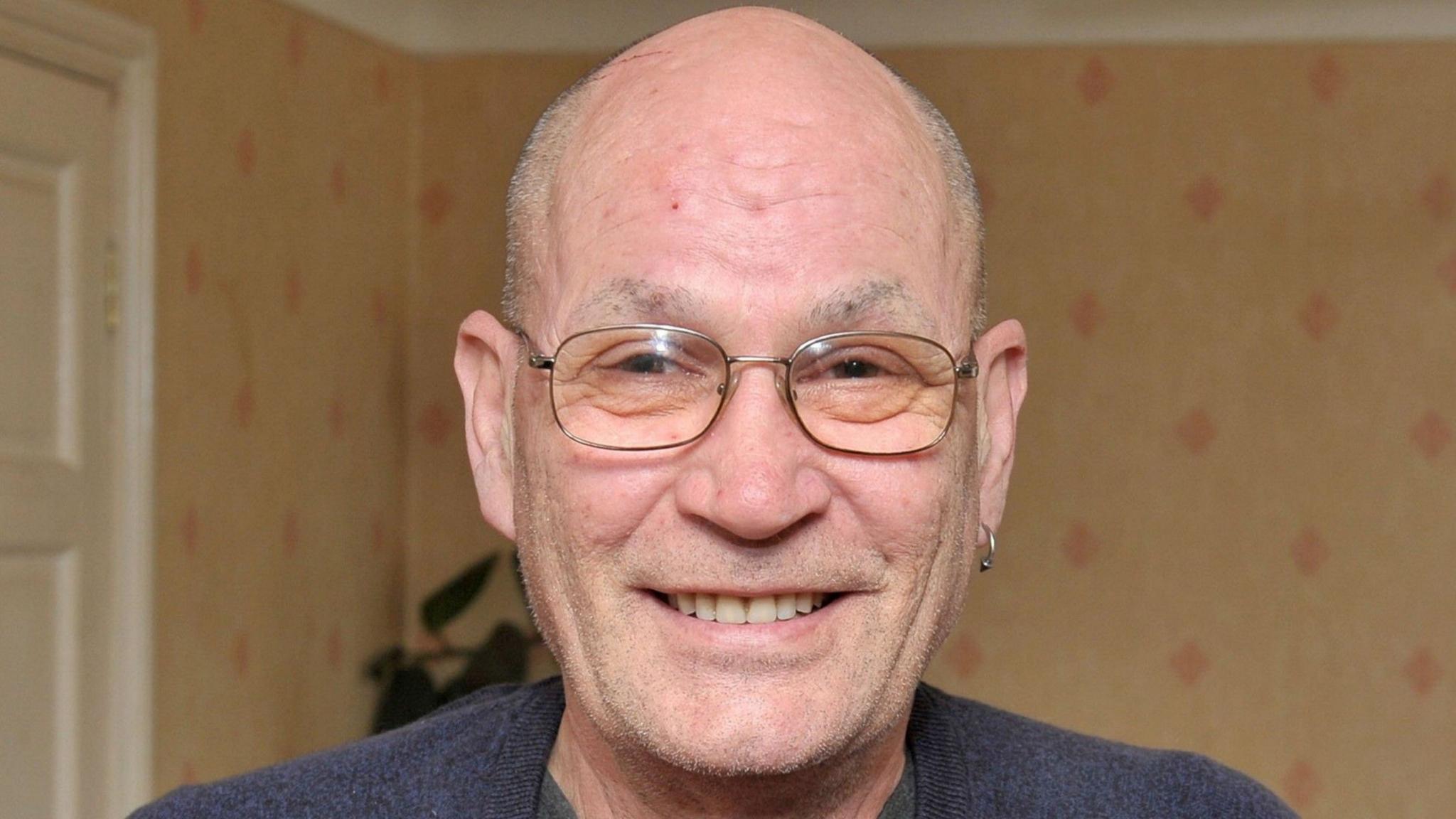 A bald man wearing a dark top and glasses. He has an earring in his left ear.