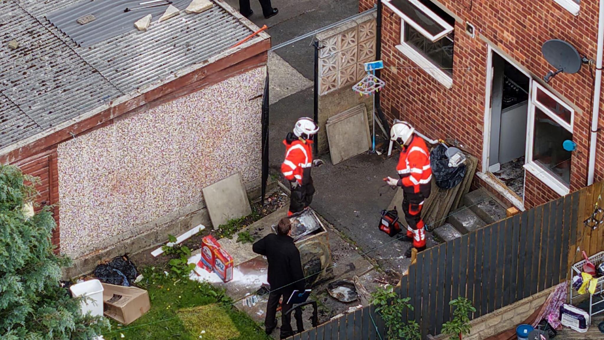 Fire investigators stand in the back garden of a semi-detached property. There is a burnt out washing machine and fire damaged items on the ground.