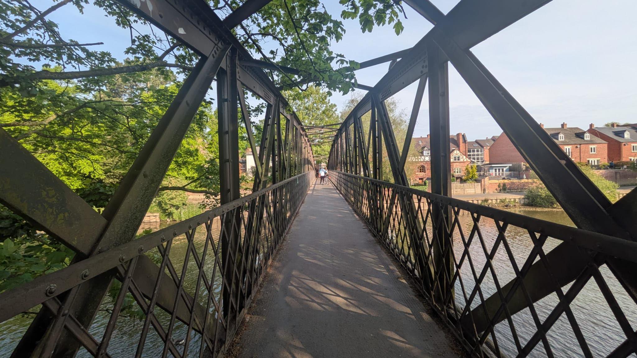 A long bridge walkway is covering a river. It is surrounded by blue sky, houses and green trees. In the long distance, we can see a person standing in the middle of the bridge.