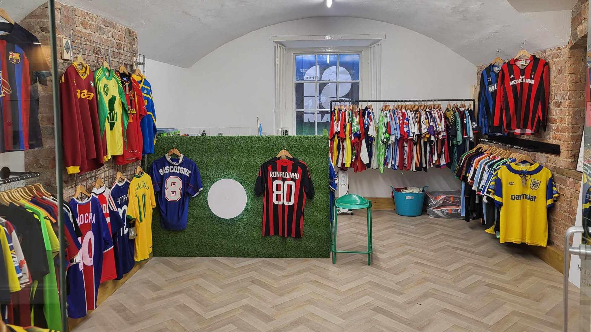 The 12th Man shop. On the left, right and back walls there are dozens of hangers holding brightly coloured retro football shirts. There is a desk that is covered in green astro turf material.