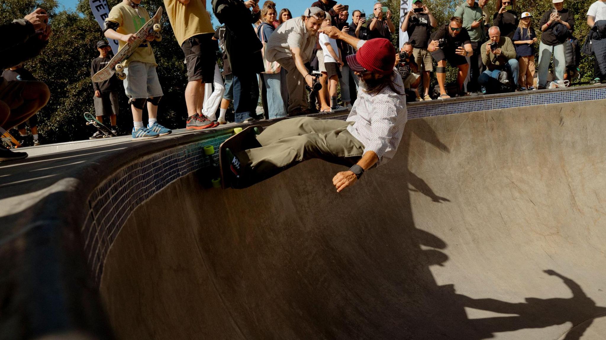 This picture shows a skateboarder performing a trick on the edge of a skate bowl. The skateboarder is dressed in a red cap, white shirt, and khaki trousers. Around the bowl, there's an audience of people—some holding cameras and skateboards—watching the action. In the background, there are trees and banners with text likely related to skateboarding.