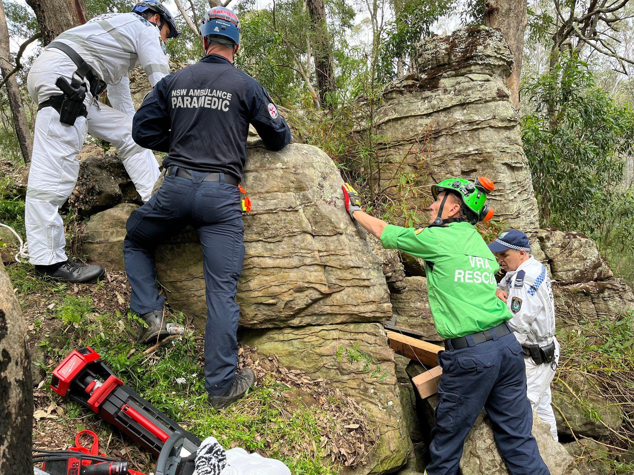 Four men - a police officer, a paramedic, a man in a white suit and another in a green top reading VAL Rescue - stand next to a large boulder in a wood. Pieces of wood used to make the rocks secure can be seen, as well as mechanical equipment