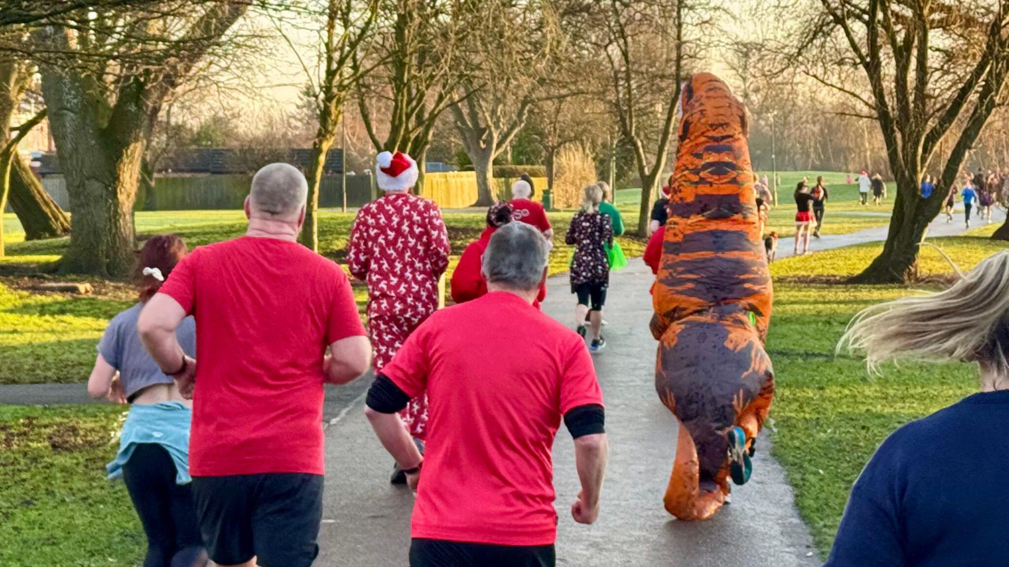 People running away from the camera on a path in a park. Someone is dressed in a large inflatable dinosaur costume.