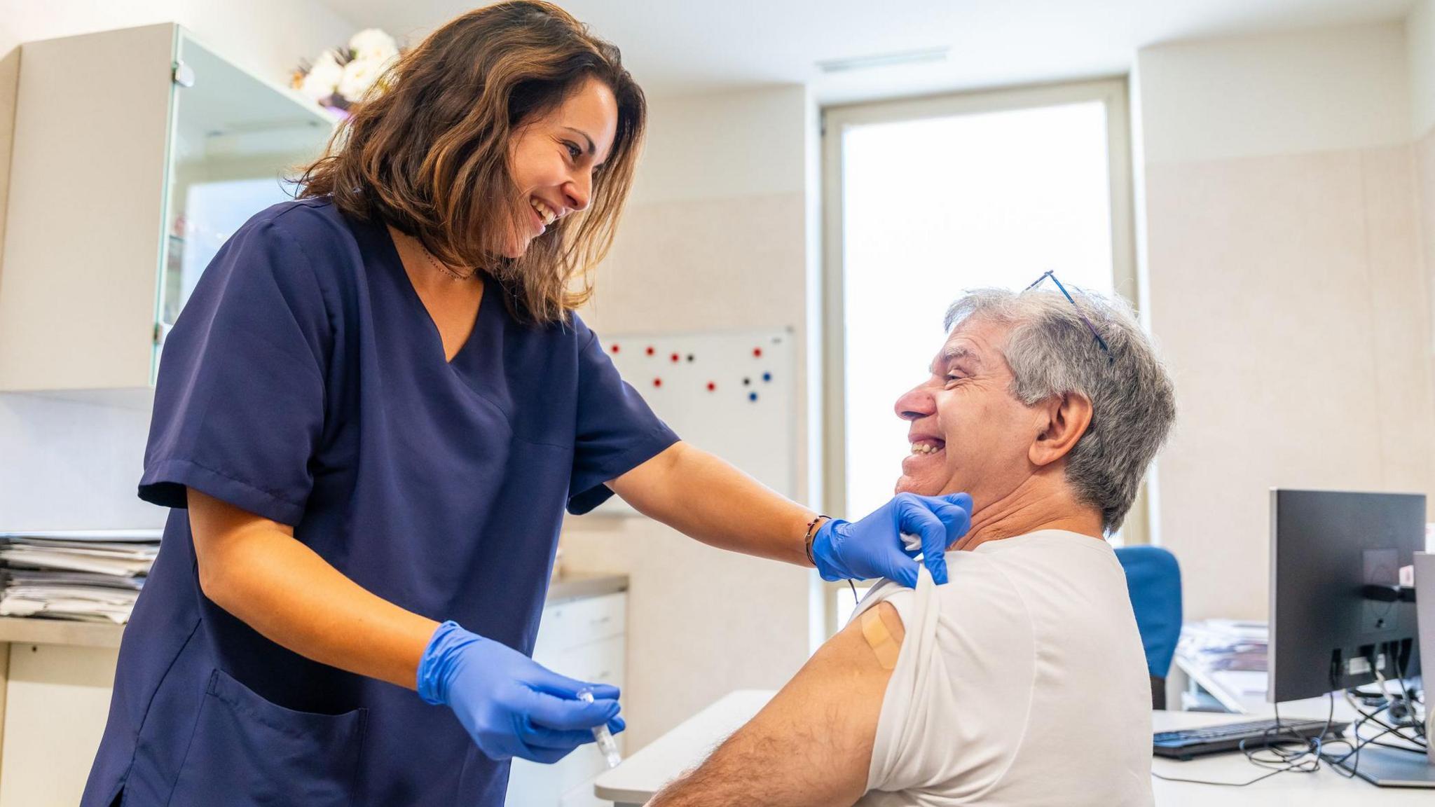 Two people in a bland, white clinic room. One is an older man with grey hair, an incredibly intense grin on his face and his left arm is rolled up. A young woman with shoulder length brown hair, wearing a rich blue set of scrubs is standing over him. She holds a shot of flu vaccine in her medical gloved hands. 