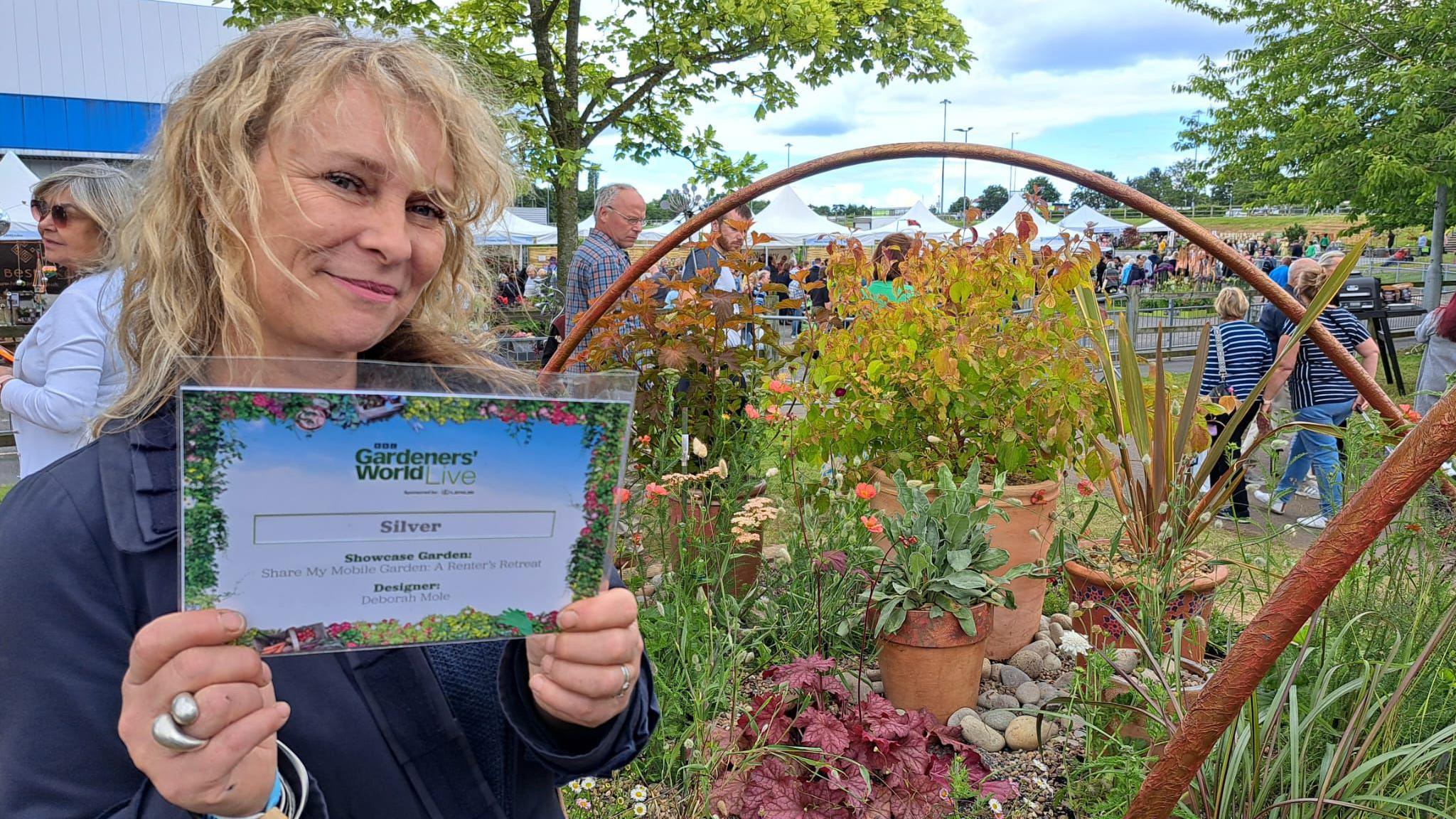 A blond woman in a navy blue coat is holding a landscaped A4 sized certificate and she is standing to the left of the picture. On the left side of the image is a decorative garden 