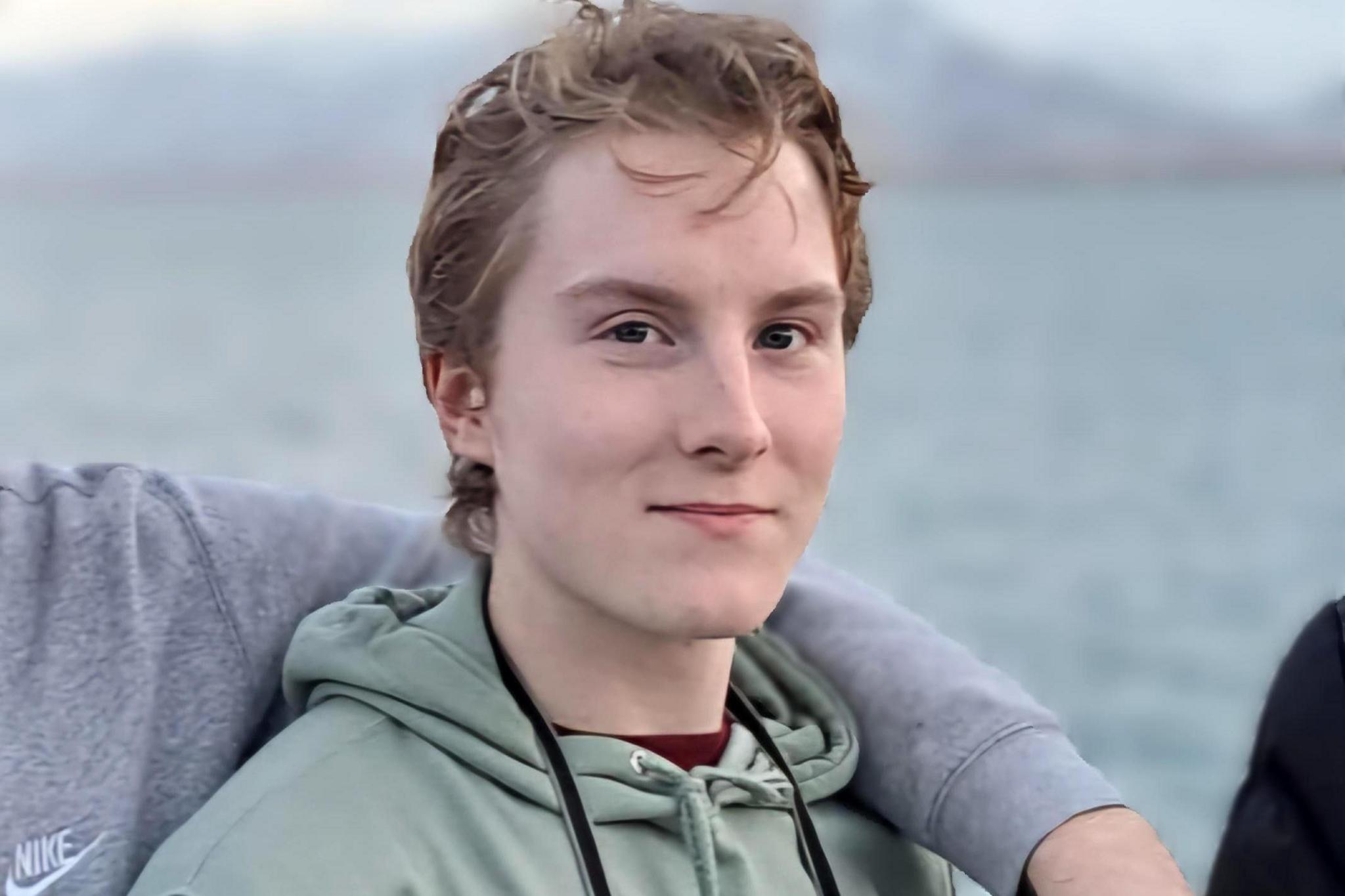 A section of a larger picture of Matthew Adams which has been cropped to show just him. An arm is resting across his back and shoulders but we can't see who it belongs to. Matthew is wearing a pale green hoodie and has the straps of a camera round his neck. He has short, strawberry-blond hair with a slightly unravelled quiff at the front. He's half smiling, as if he's familiar with the person taking the photograph.