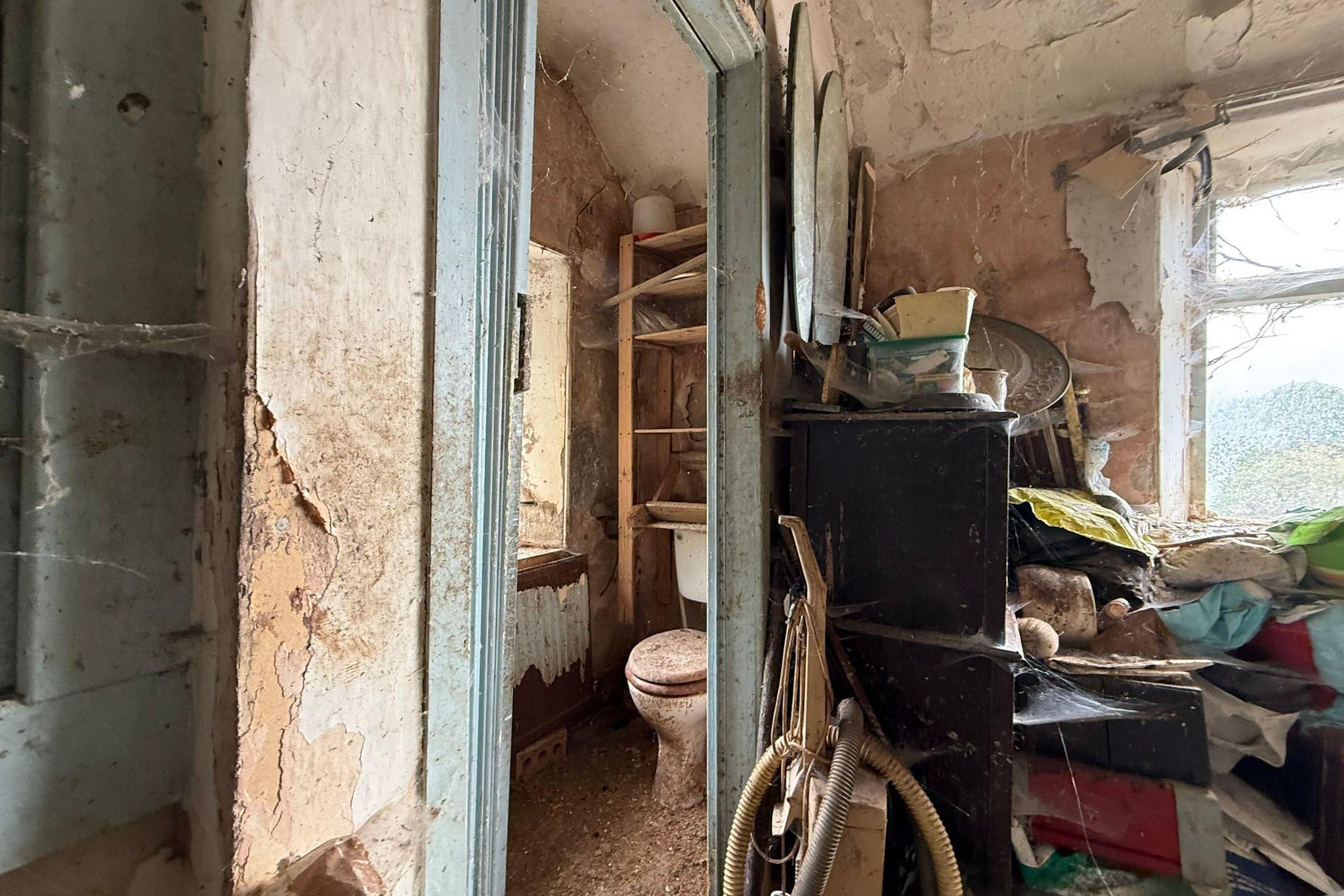 One of the bathrooms which is covered in dirt and stains. The picture has been taken from just outside the bathroom. On the left, there is a pile of junk just below the window which is covered in cobwebs. 