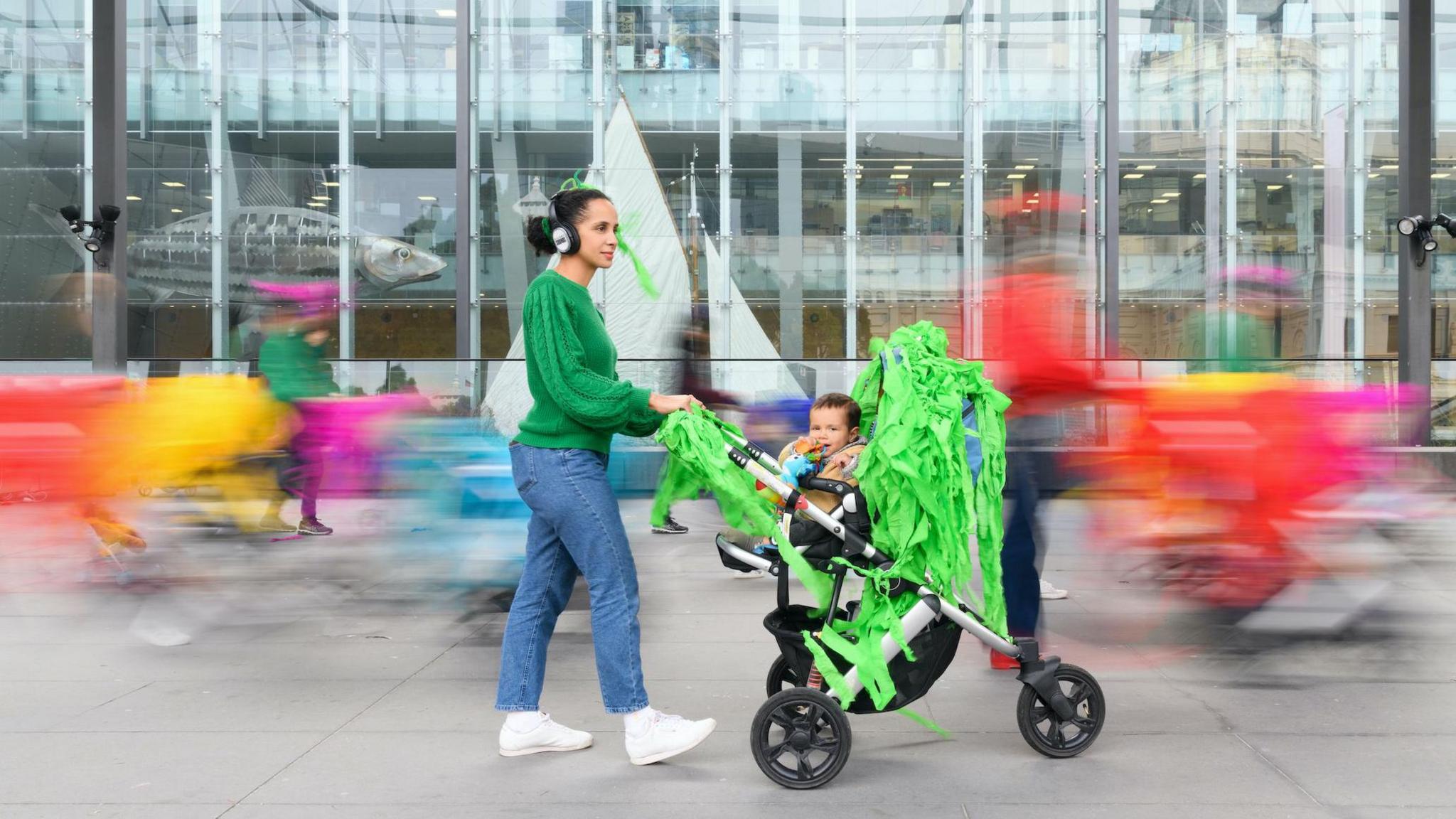 Shot of a woman wearing headphones, a green top and jeans pushing a pram decorated with bright-green material with blurred coloured-shapes in the background